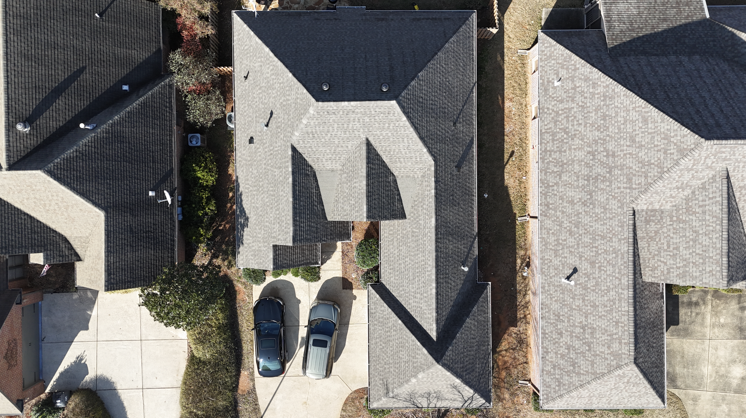 Aerial view of several houses with gray shingle roofs, driveways, trees, and parked cars.