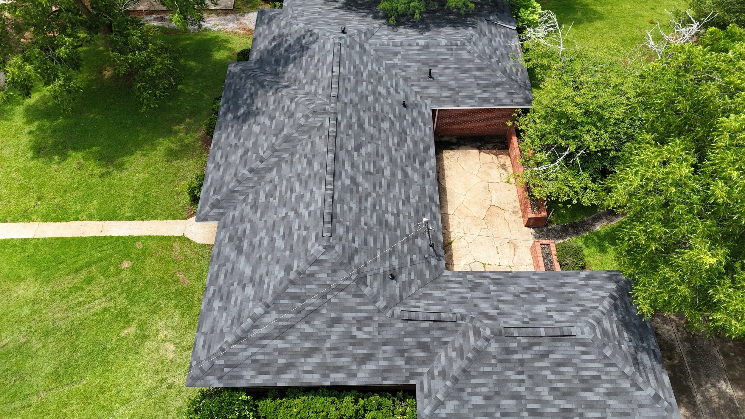An aerial view of a house with a gray shingled roof, a concrete driveway, and surrounded by green trees and grass.