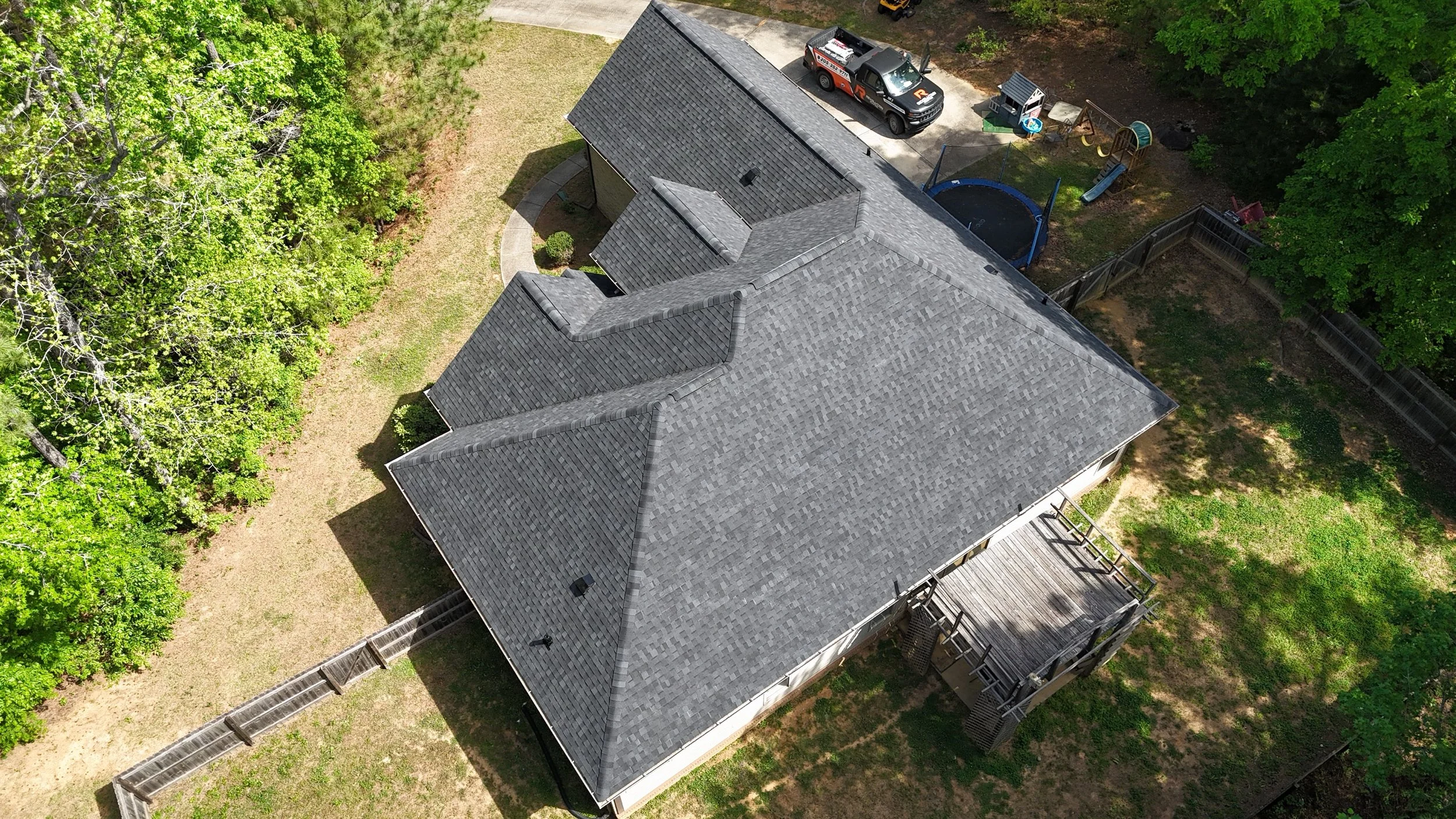 An aerial view of a house with gray shingle roof, surrounded by green trees, a fenced backyard, a driveway with a pickup truck, and a small children's play area.