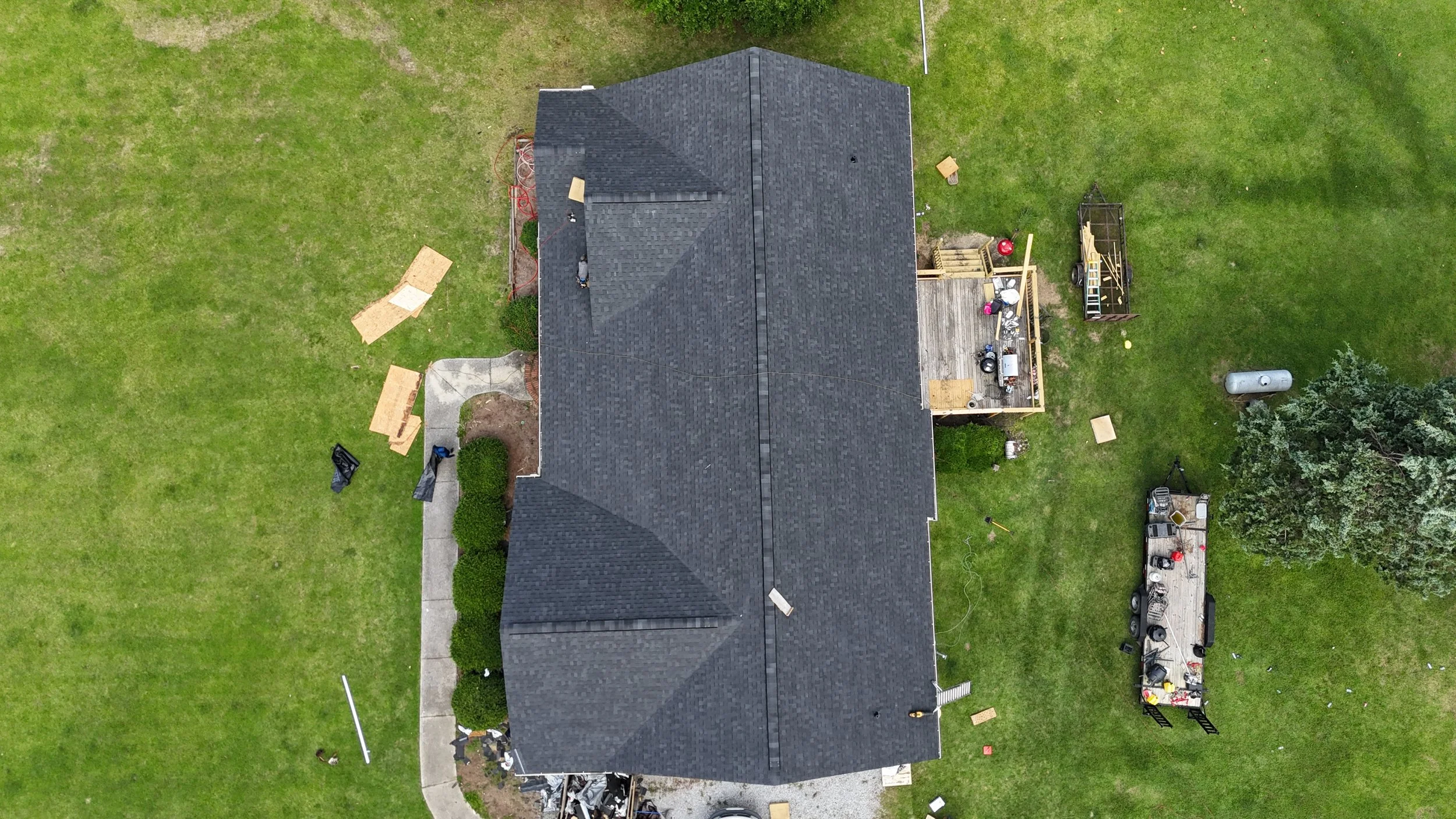 An aerial view of a house with a black roof, surrounded by green grass. The backyard has construction items, a small deck, and a trailer with tools and equipment.