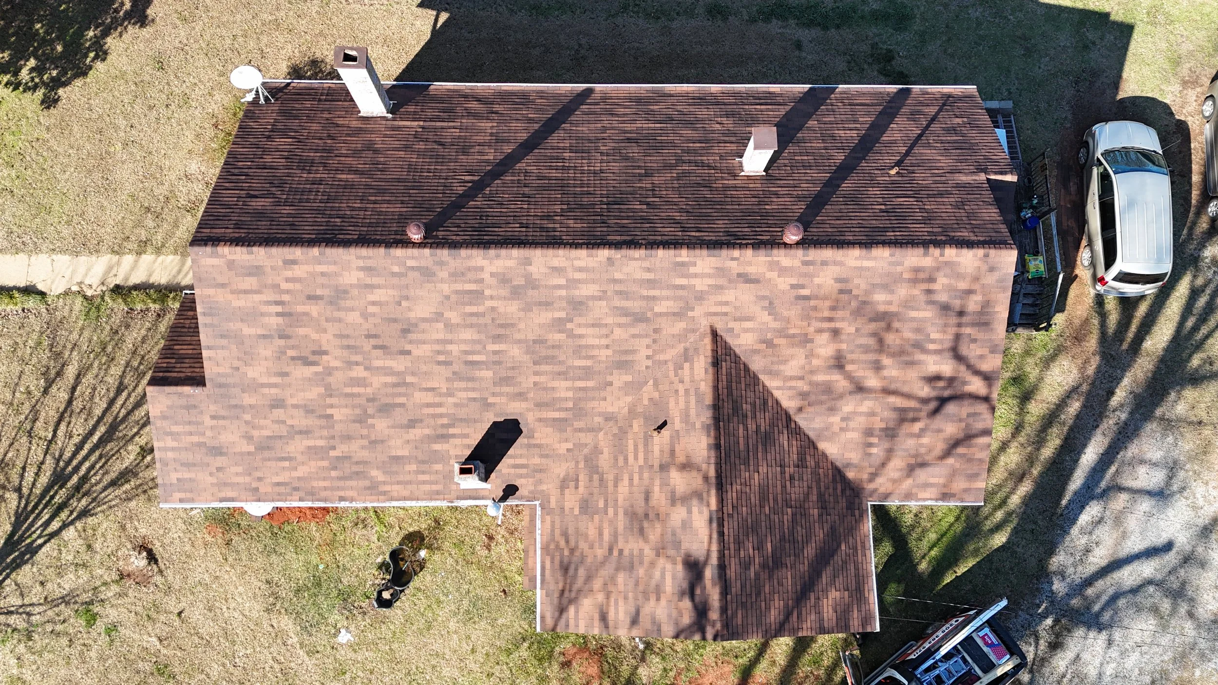 An aerial view of a house with a brown shingled roof, chimney, and vents. There is a driveway with a silver SUV parked on the side, and a trailer with tools near it. The yard has grass, trees, and a concrete walkway.