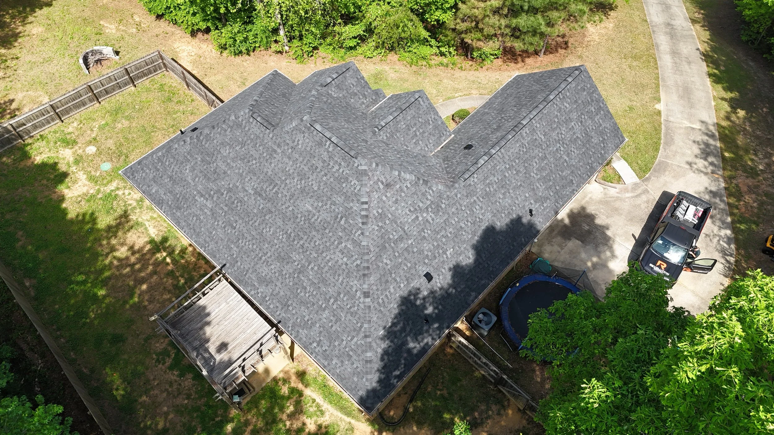 Aerial view of a house with a gray shingle roof, surrounded by green trees, a wooden fence, and a curved driveway with a pickup truck parked.