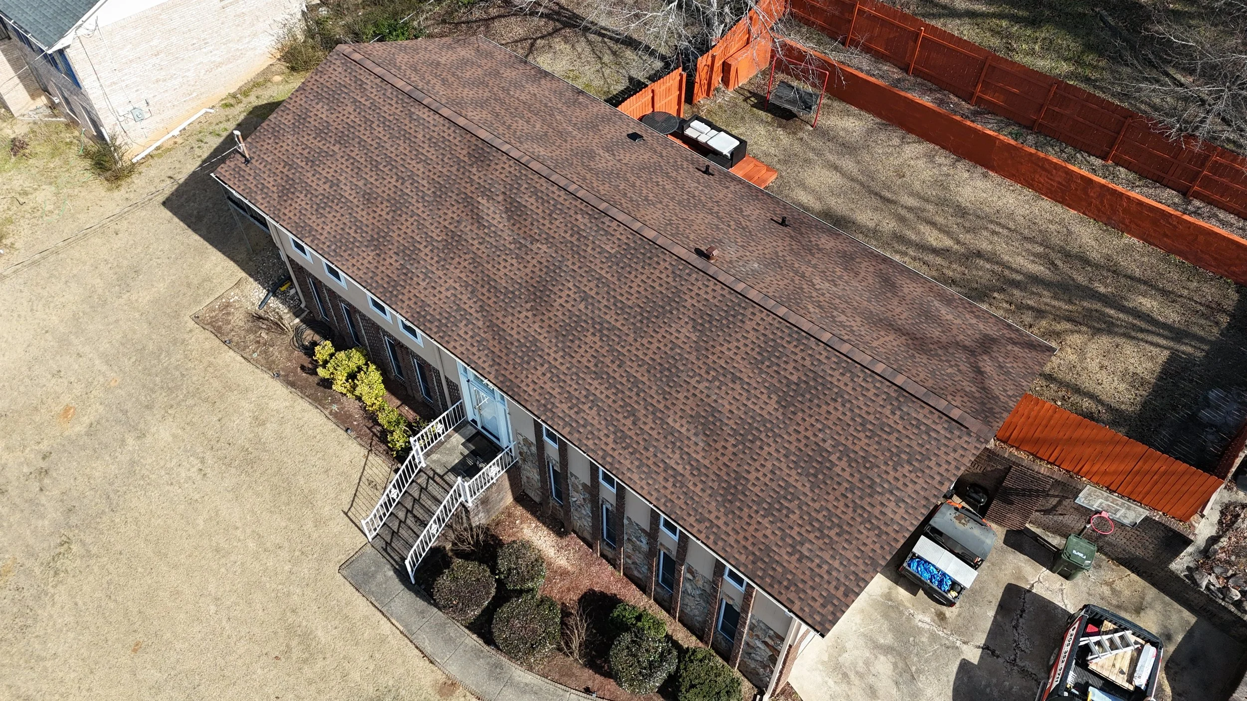 Aerial view of a multi-unit residential building with a brown tiled roof, beige exterior walls, and white window frames. The building has small landscaped areas with shrubs, and is surrounded by gravel and dirt yards with a red fence on one side. There are vehicles parked near the building, and a backyard with a trampoline and patio furniture.