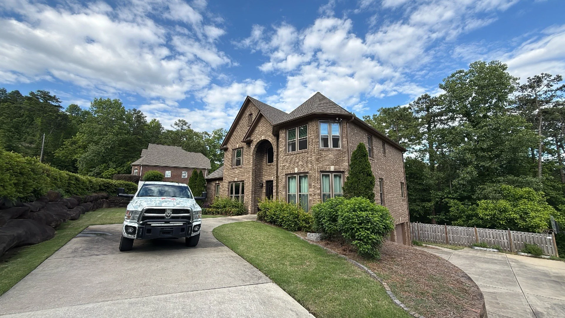 A brick house with a driveway, a white truck parked in front, surrounded by green trees and bushes, under a blue sky with clouds.