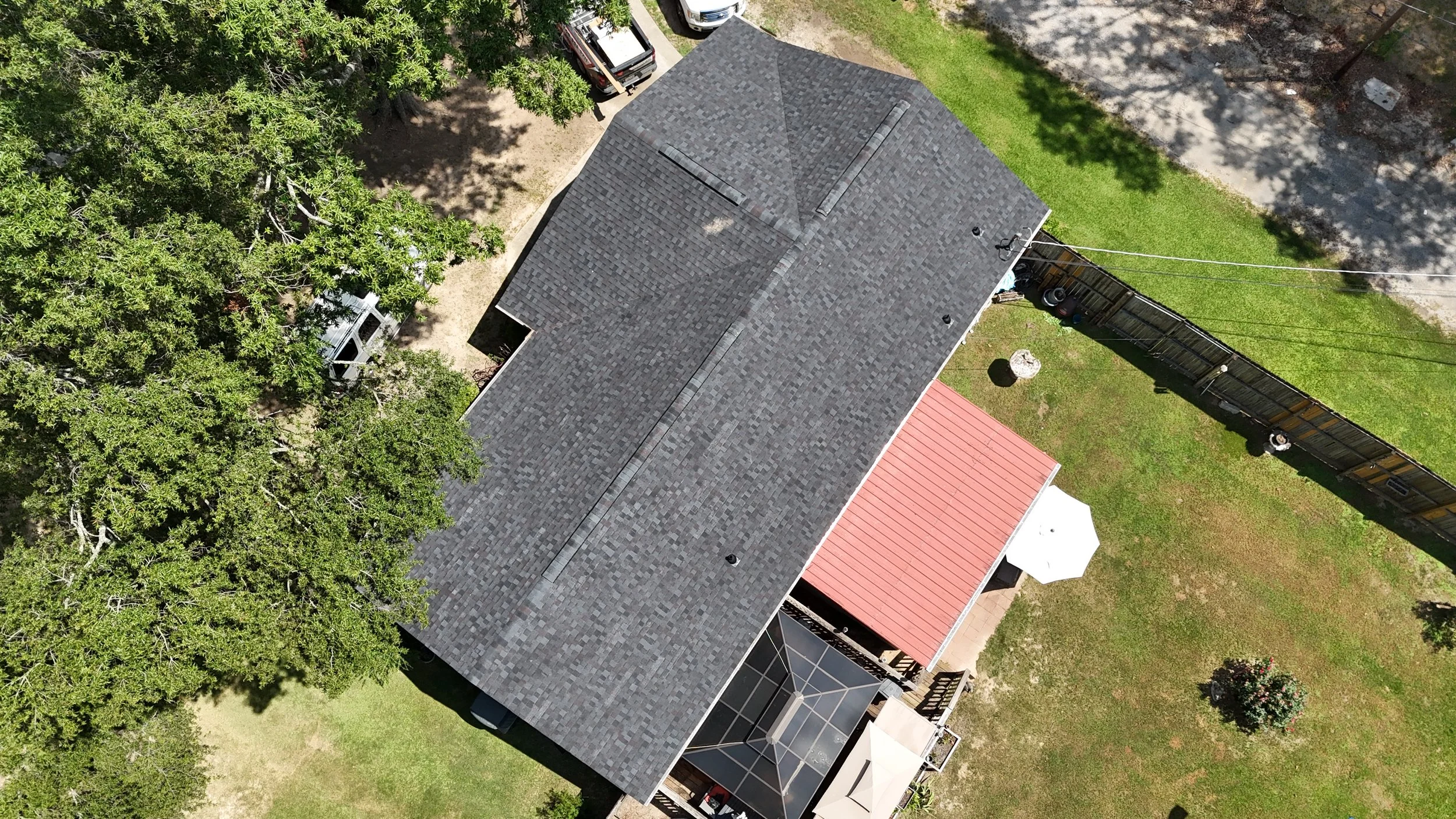 An aerial view of a house with a gray shingle roof, surrounded by green trees, a backyard with a red roof outdoor seating area, and a wooden fence, with vehicles parked nearby.