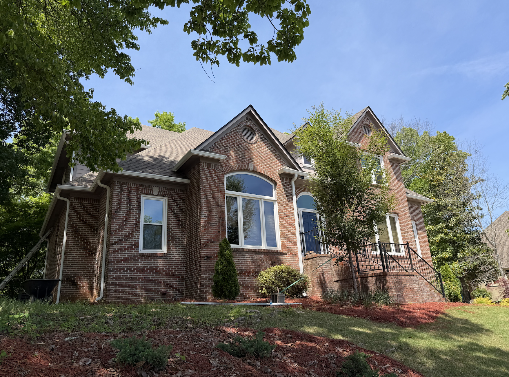 A large, elegant brick house with multiple gables and large front windows, surrounded by mature trees and a well-maintained yard with newly planted landscaping.