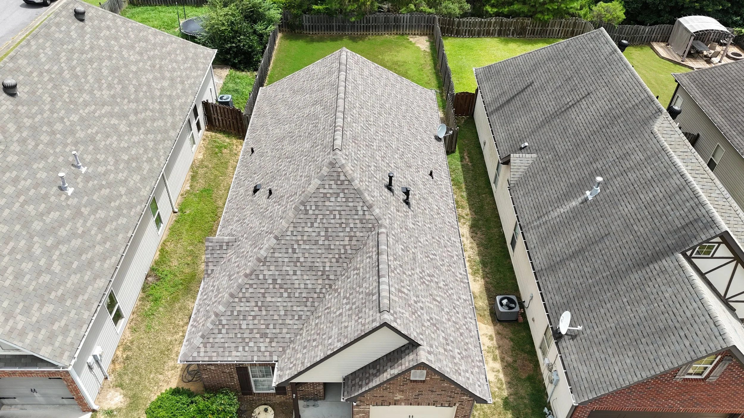 An aerial view of three neighboring houses with tiled roofs, small yards, and fenced backyards, with a grassy area and backyard structures visible.
