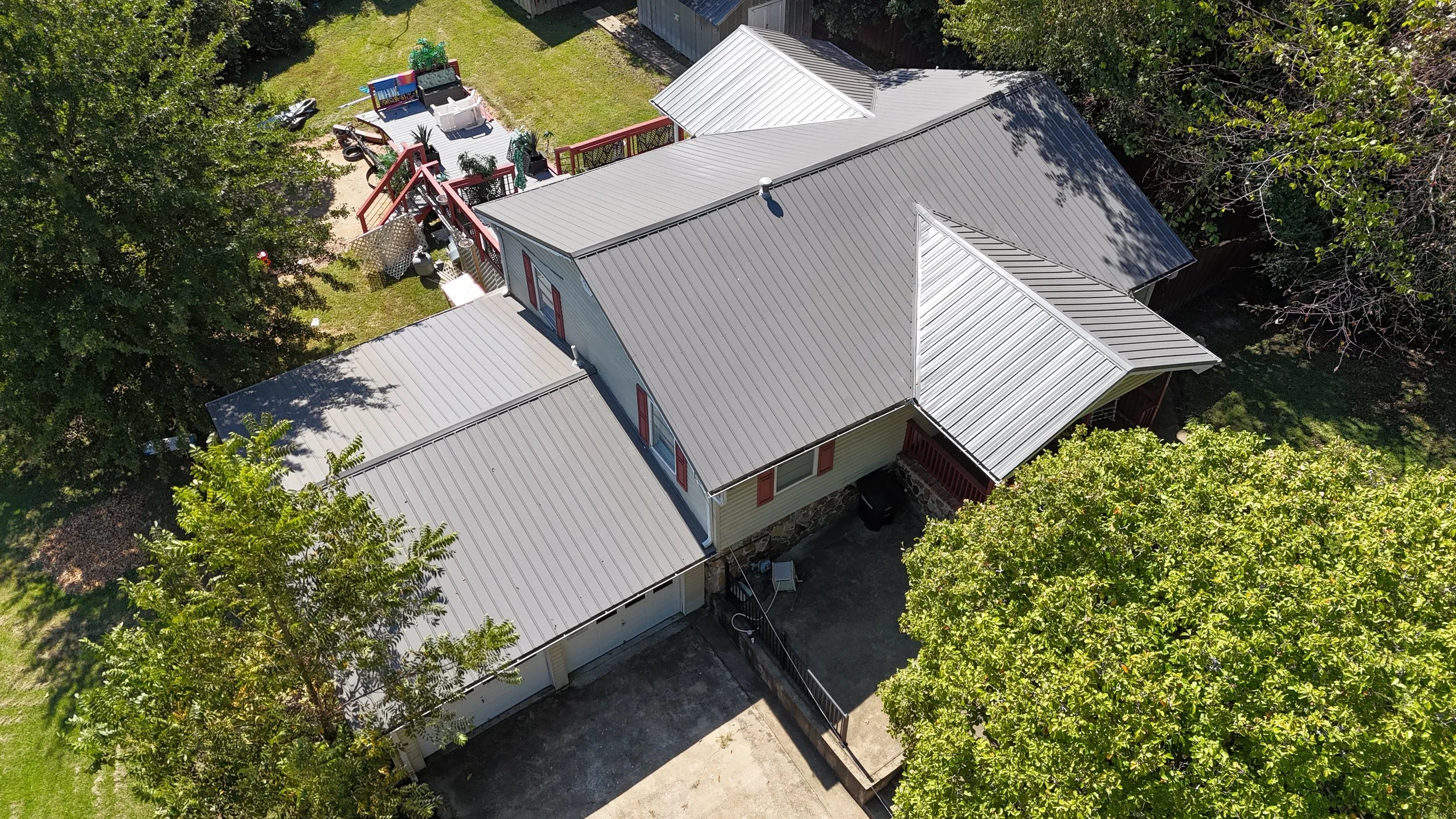 An aerial view of a house with a metal roof, surrounded by trees and a fenced yard with outdoor furniture and toys.