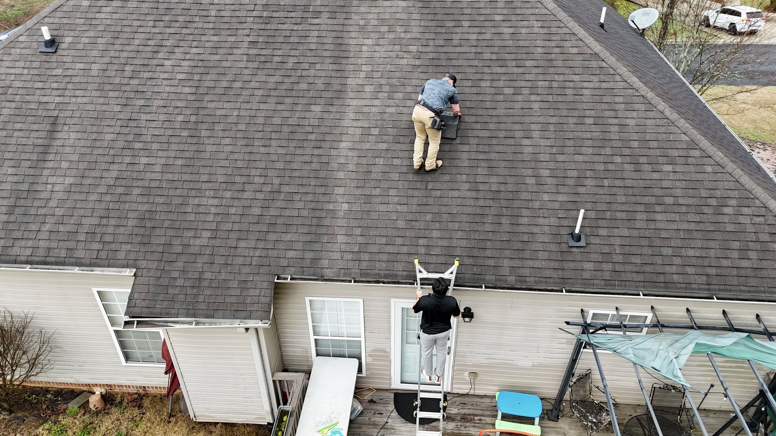Two people on the roof of a house, one working near the chimney and the other observing, with a ladder on the deck below.