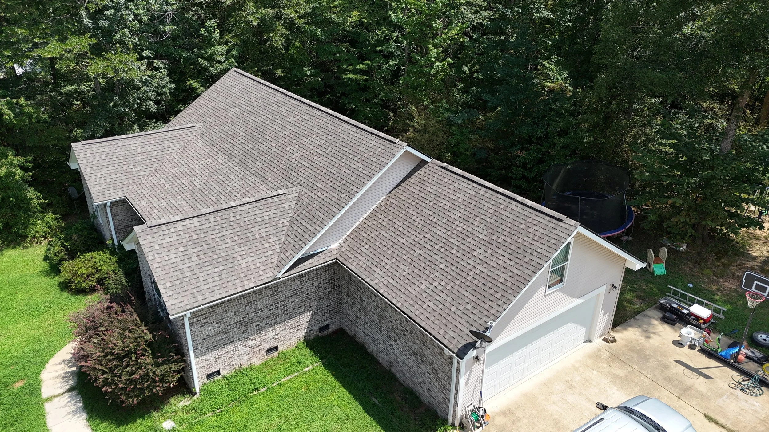 An aerial view of a house with a gray shingle roof, brick walls, and a large backyard surrounded by trees. There is a trampoline, a basketball hoop, a small shed, and some children’s toys visible in the yard.