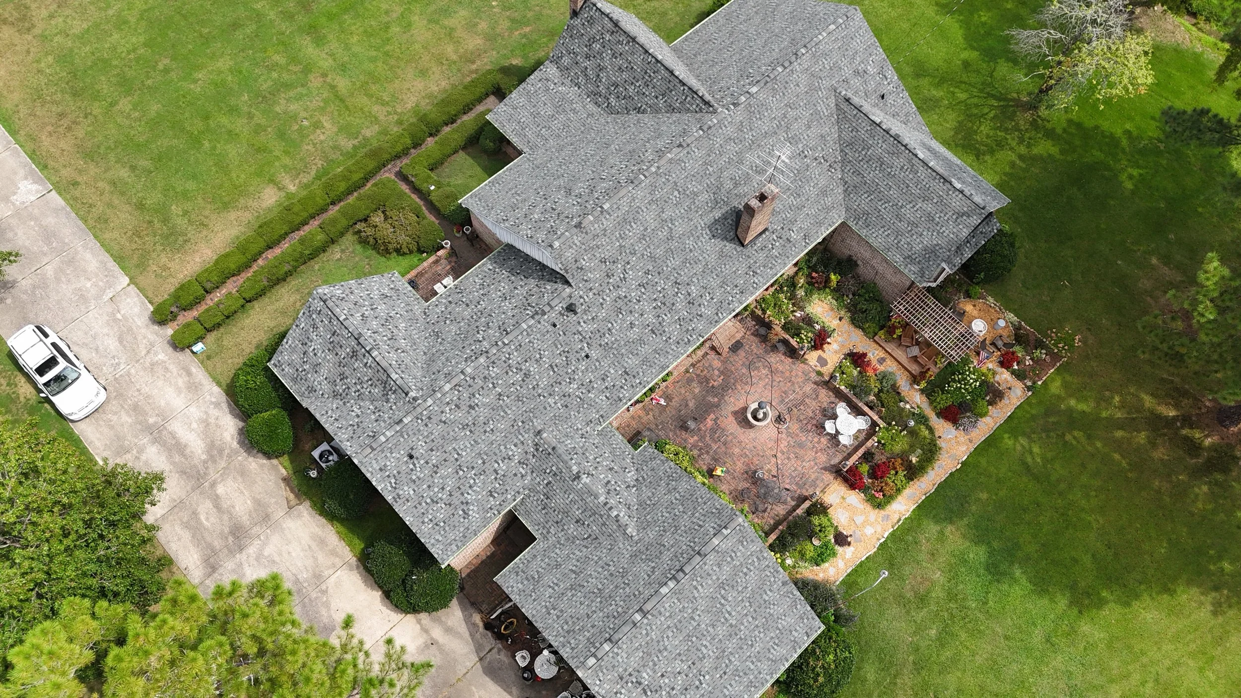 An aerial view of a house with a gray shingle roof, a brick chimney, and surrounding landscaped yard with trees, bushes, a patio with outdoor furniture, a driveway, and a lawn.