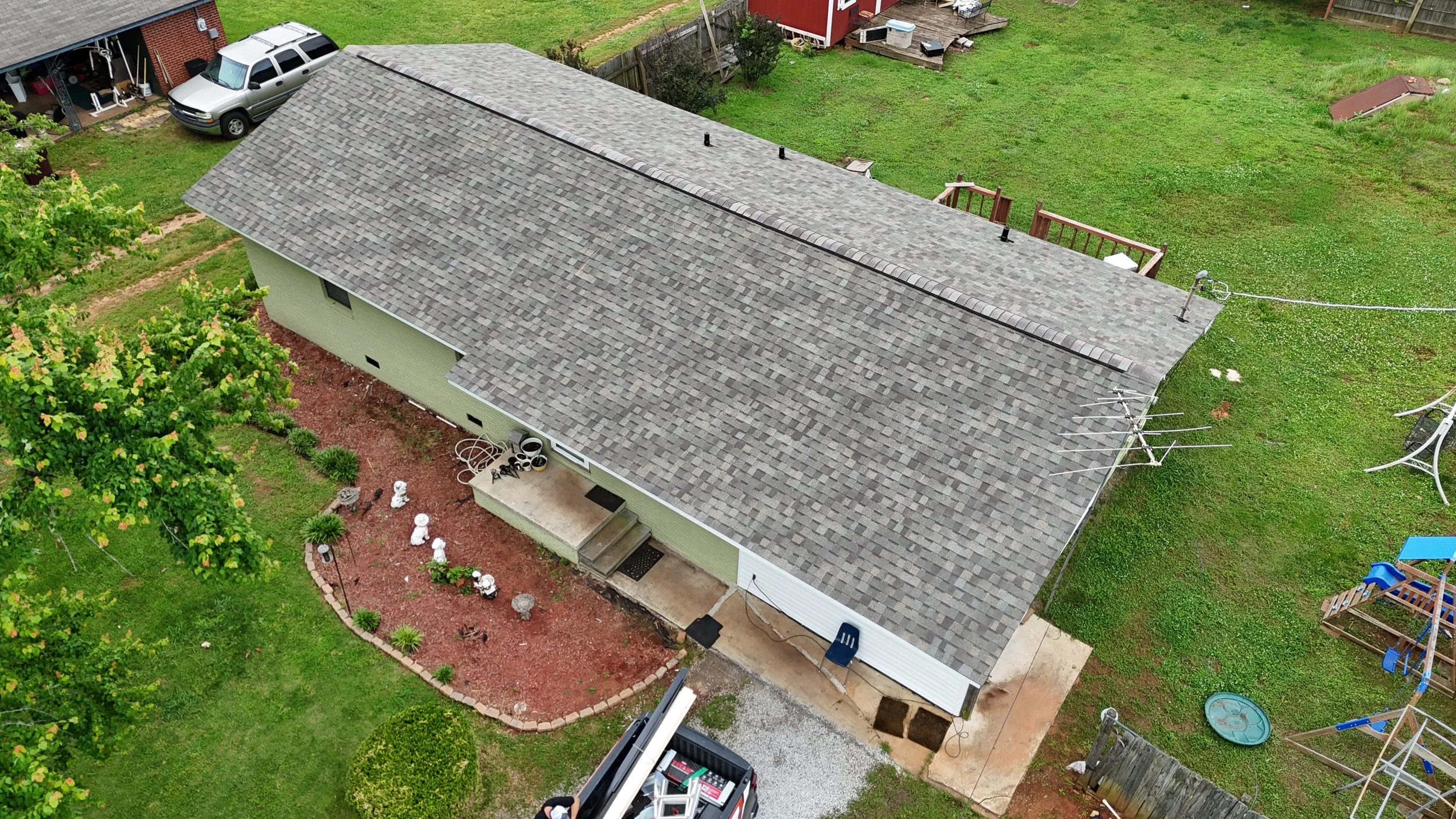 Aerial view of a house with a gray shingled roof, green exterior walls, and a backyard with a red brick garden bed, trees, and outdoor items like a blue slide, swings, and yard decorations.