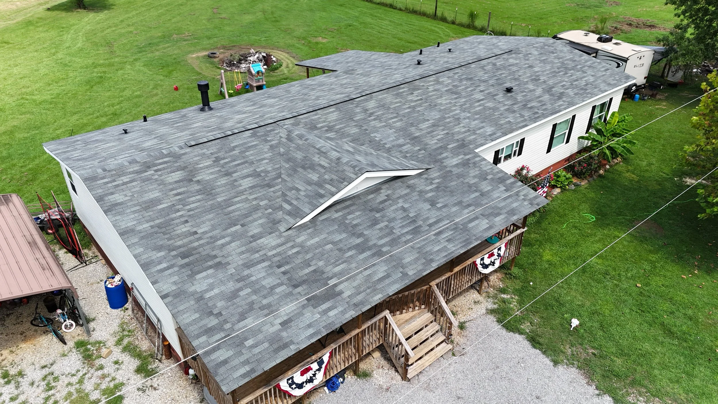 Aerial view of a house with a dark gray shingle roof, surrounded by green grass and trees, with a small backyard play area and a RV parked nearby.