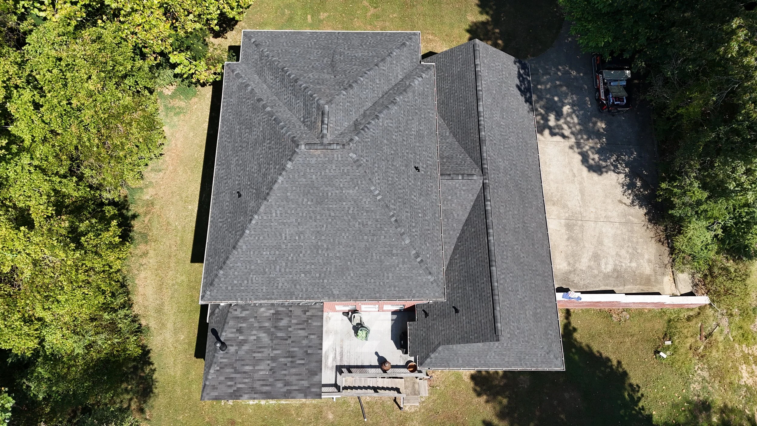 Aerial view of a house with a gray shingled roof, surrounded by green trees and a small yard, with a driveway and garage on the right side.