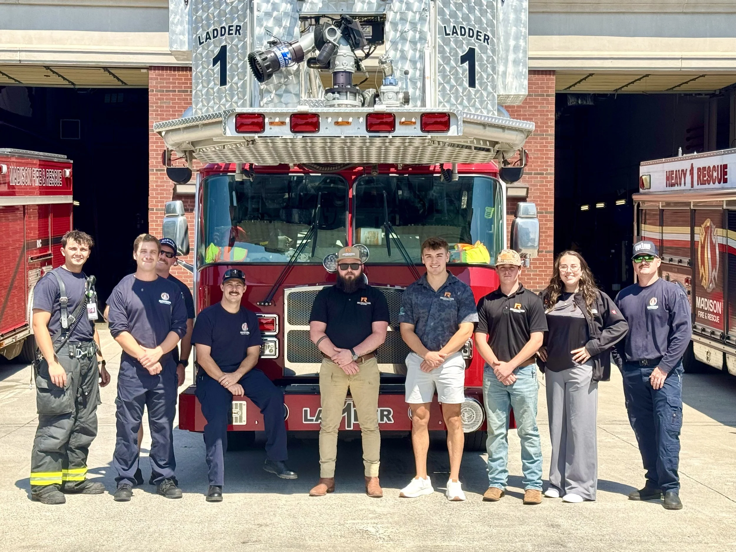 A group of nine people standing in front of a red fire truck and a fire station, some wearing firefighting gear and others in casual clothing, smiling for the photo.