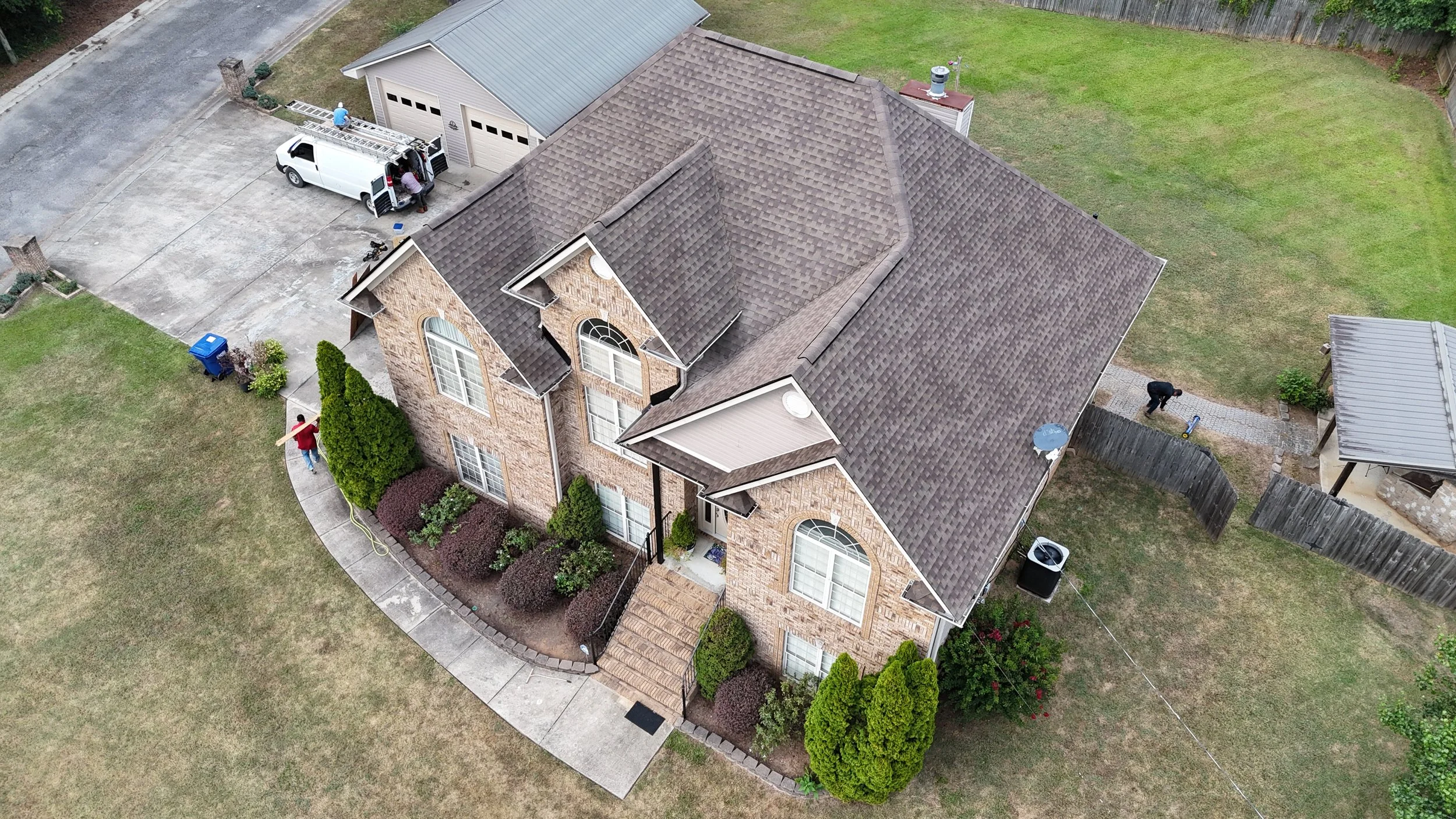 An aerial view of a brick house with a brown shingled roof, surrounded by a lawn, sidewalk, and fence. A person is carrying a wooden board near the entrance, and two workers are on a ladder on a white van parked in the driveway.
