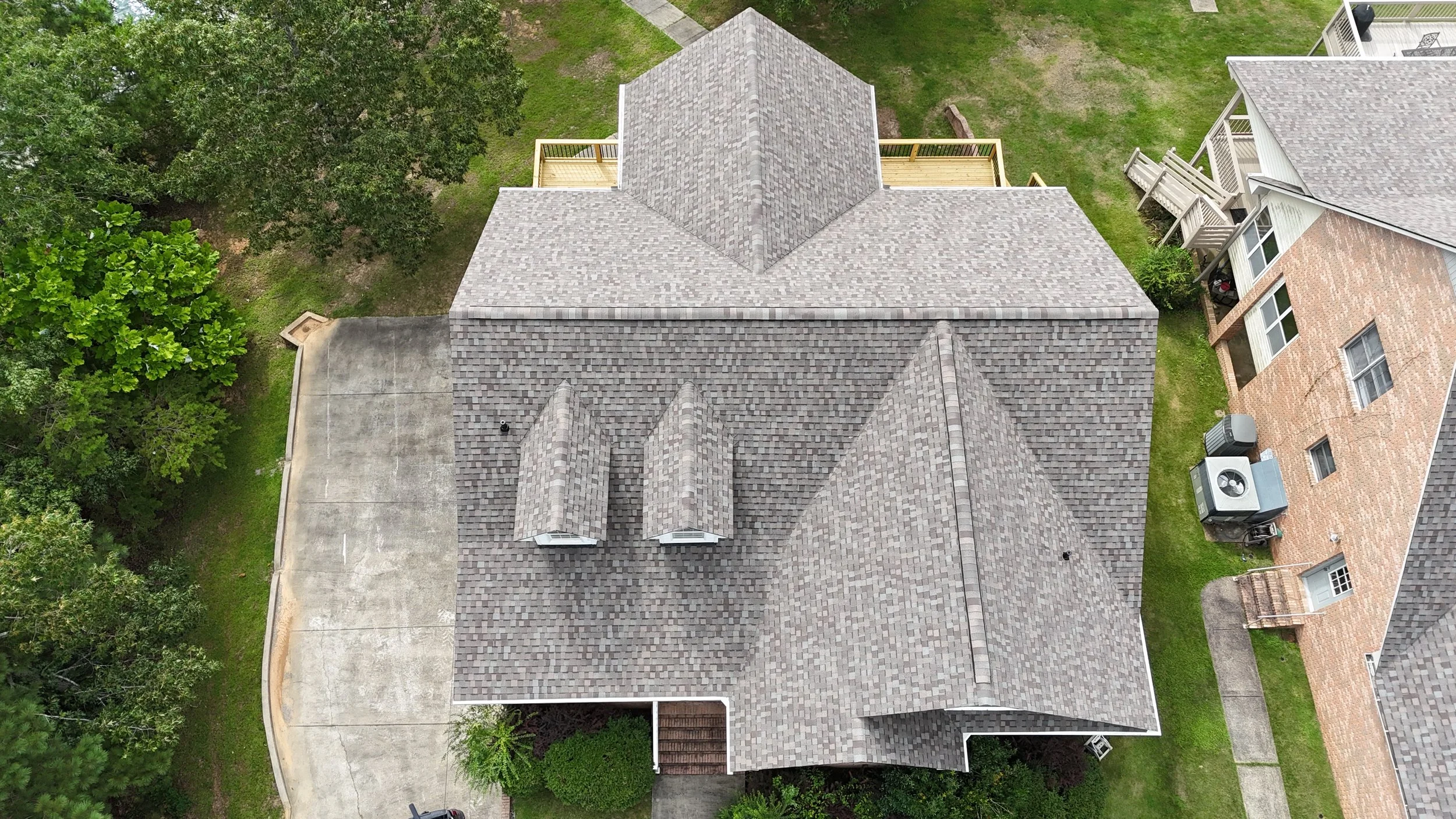 Aerial view of a house with a gray shingle roof, surrounded by green trees, a concrete driveway, and neighboring houses with brick and siding exteriors.