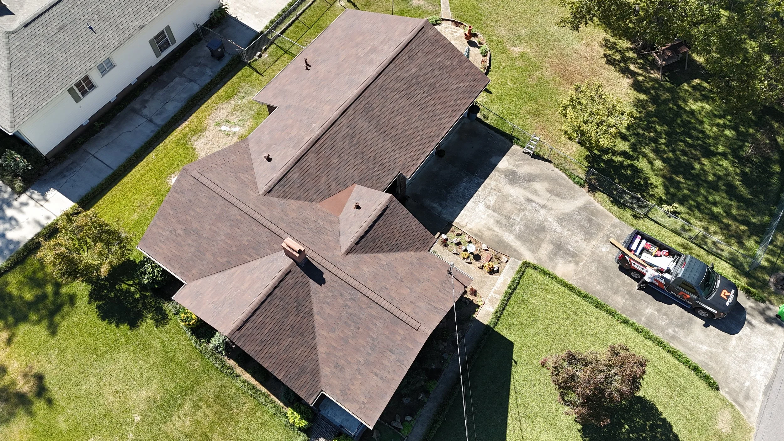 Aerial view of a house with a brown roof, driveway, and surrounding green lawn and trees. A red and white utility truck is parked on the driveway.