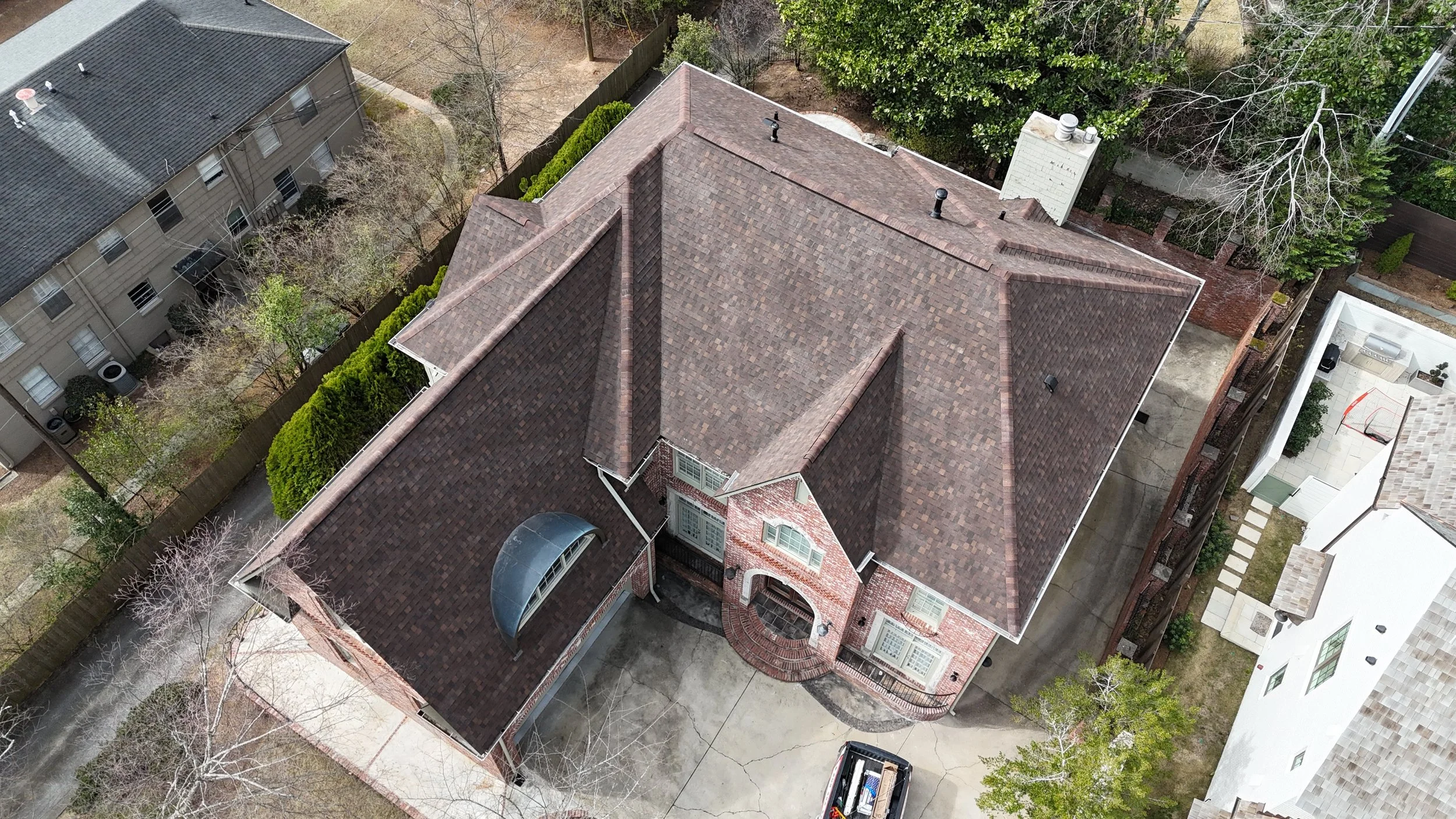 Aerial view of a large house with a brown shingle roof, brick walls, and a curved front porch. Surrounding the house are trees, a driveway with a car, and neighboring buildings, with some trees having bare branches.