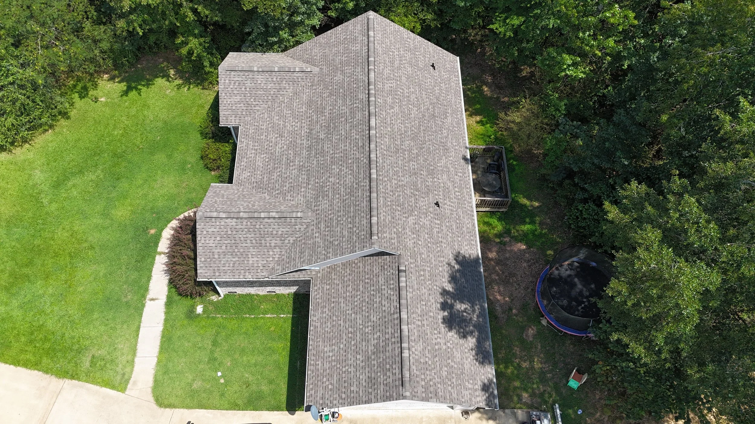 An aerial view of a house with a gray shingled roof, surrounded by green grass, trees, and a trampoline in the backyard.