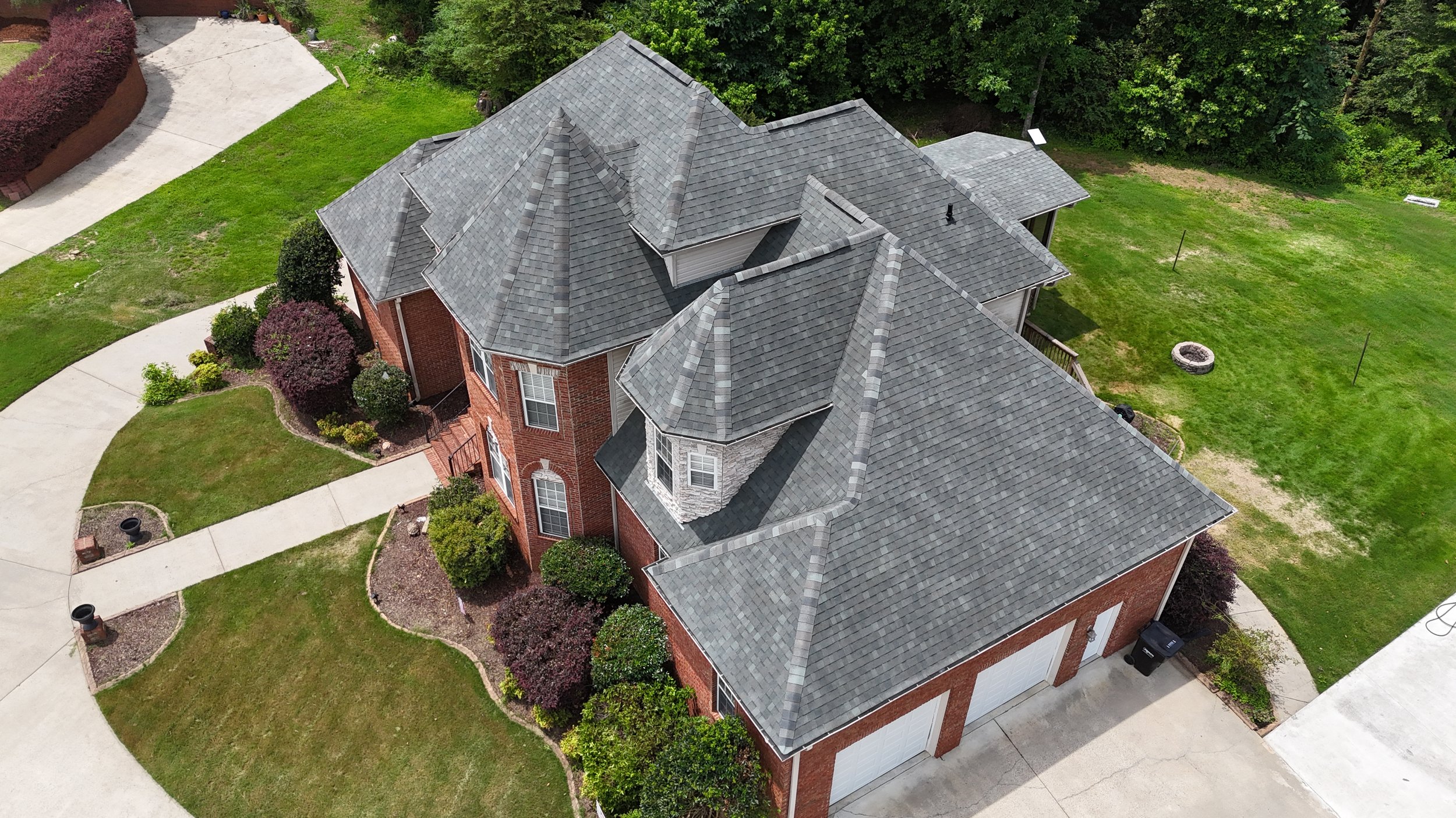 Aerial view of a two-story brick house with multiple gray shingled roof sections, surrounded by green lawn, trees, and a driveway.