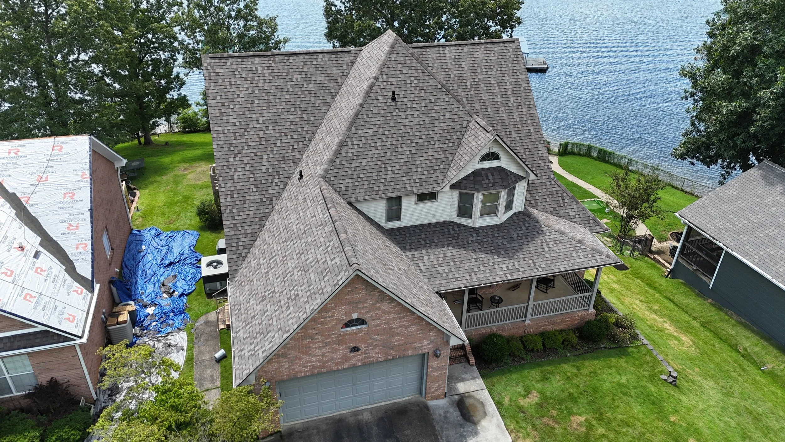 An aerial view of a house with new construction on the left side, showing partially installed roofing materials, and an existing house with a completed roof on the right, beside a body of water with a dock. The yard features green grass, trees, and a walkway.