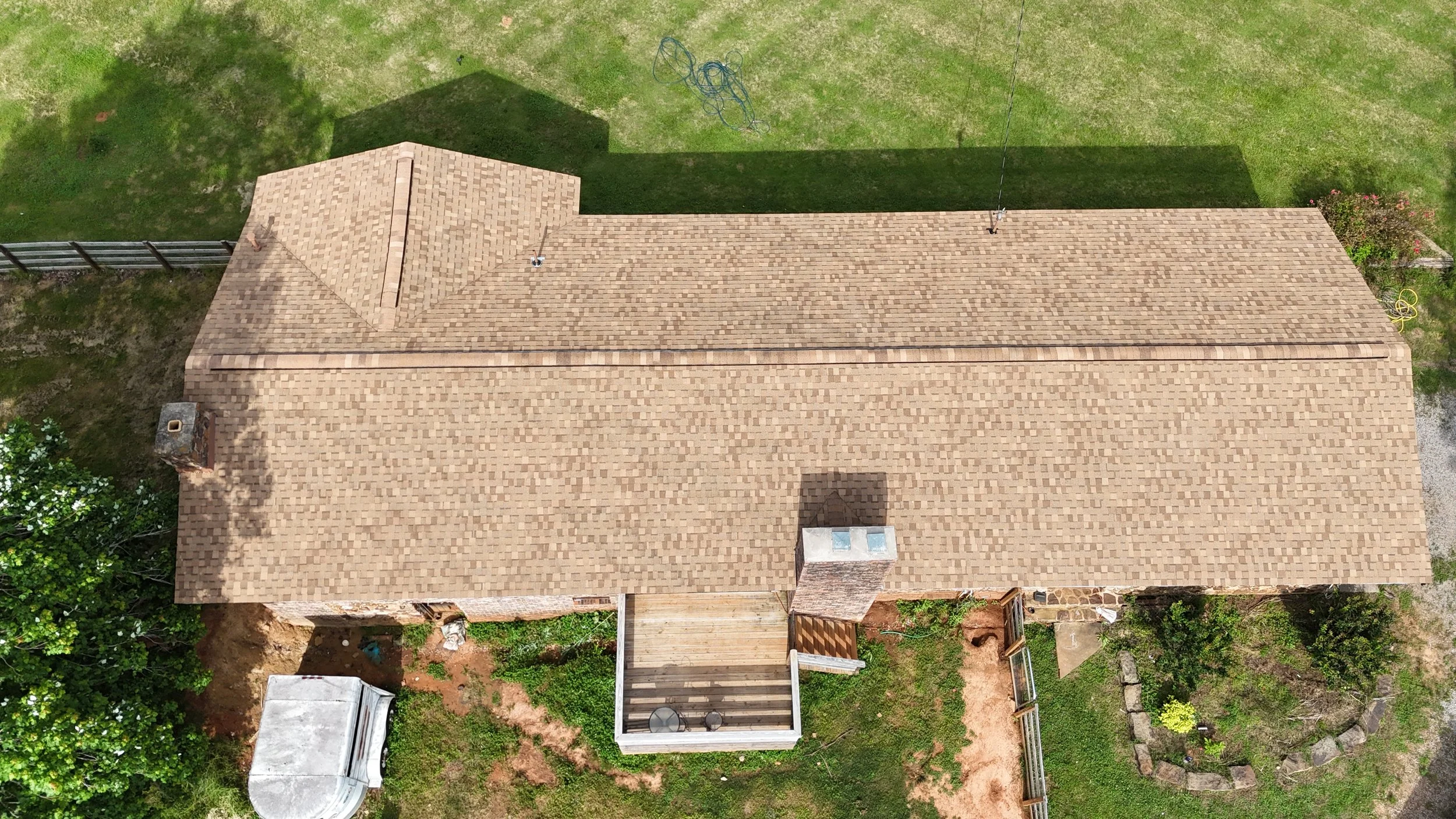 Aerial view of a house with a tan-colored shingled roof, a small outdoor deck, and green grass surrounding it.