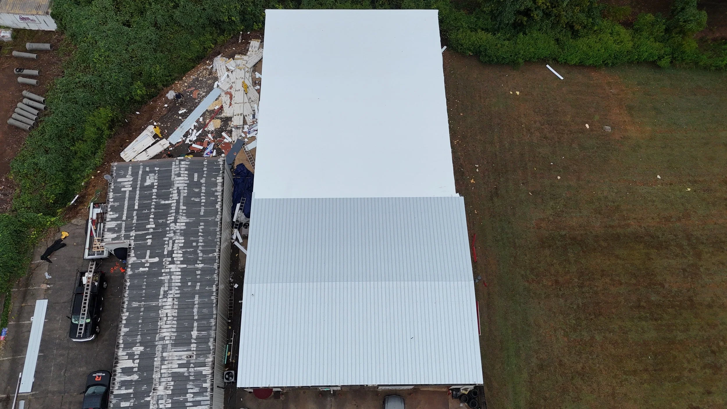 An aerial view of a construction site showing a partially built building with a black roof, a white roof, and construction materials scattered around. There are vehicles parked nearby and greenery surrounding the site.