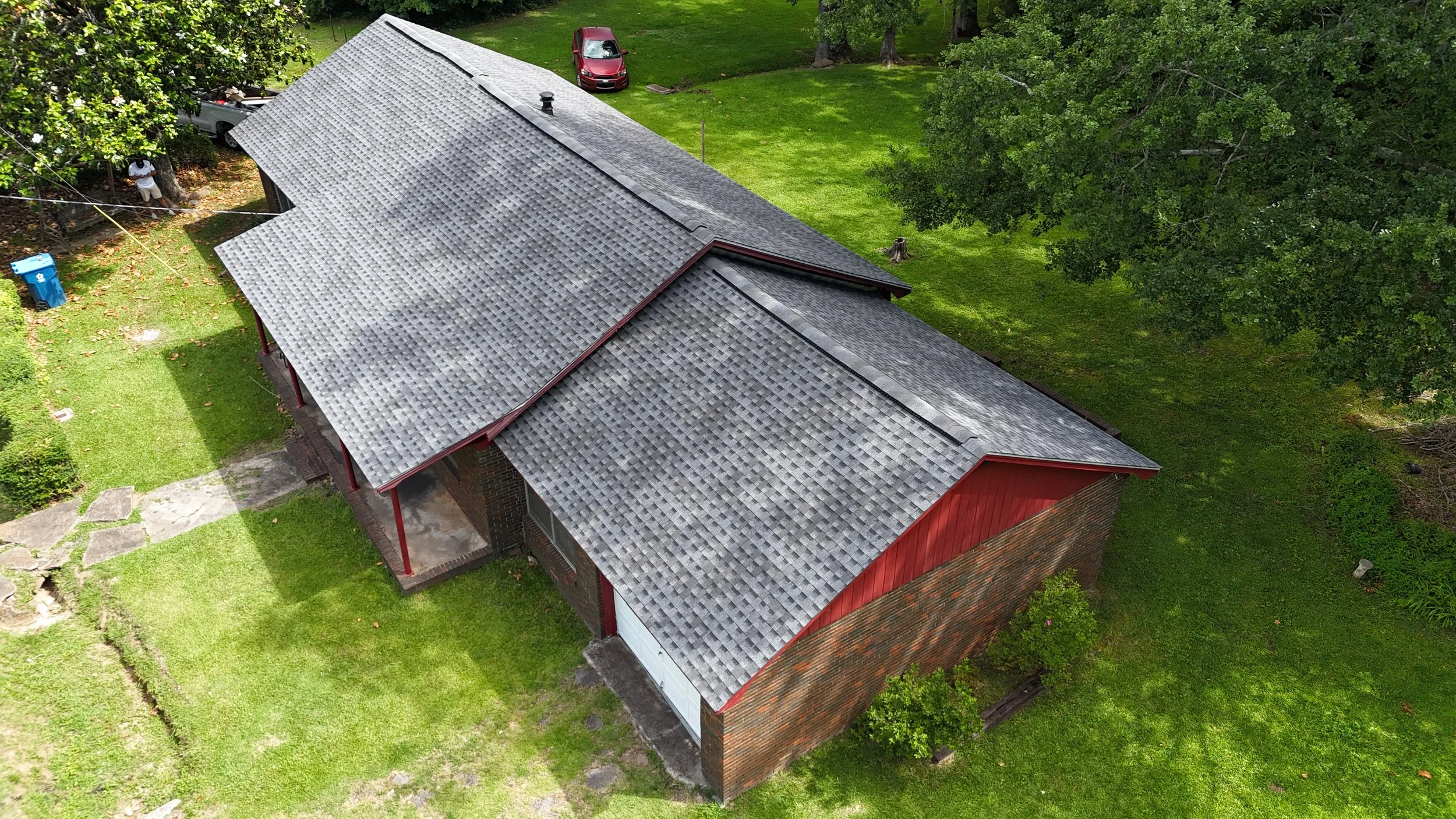 Aerial view of a house with a gray shingled roof surrounded by green lawn and trees.