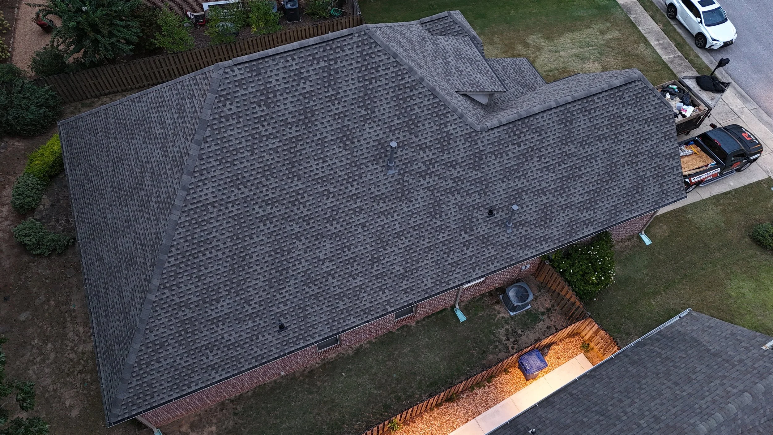 An aerial view of a house with a dark gray shingle roof, a small window, and a chimney. There is a driveway with a white car and a black pickup truck parked, and a backyard with a wooden fence, trees, bushes, and some garden beds.