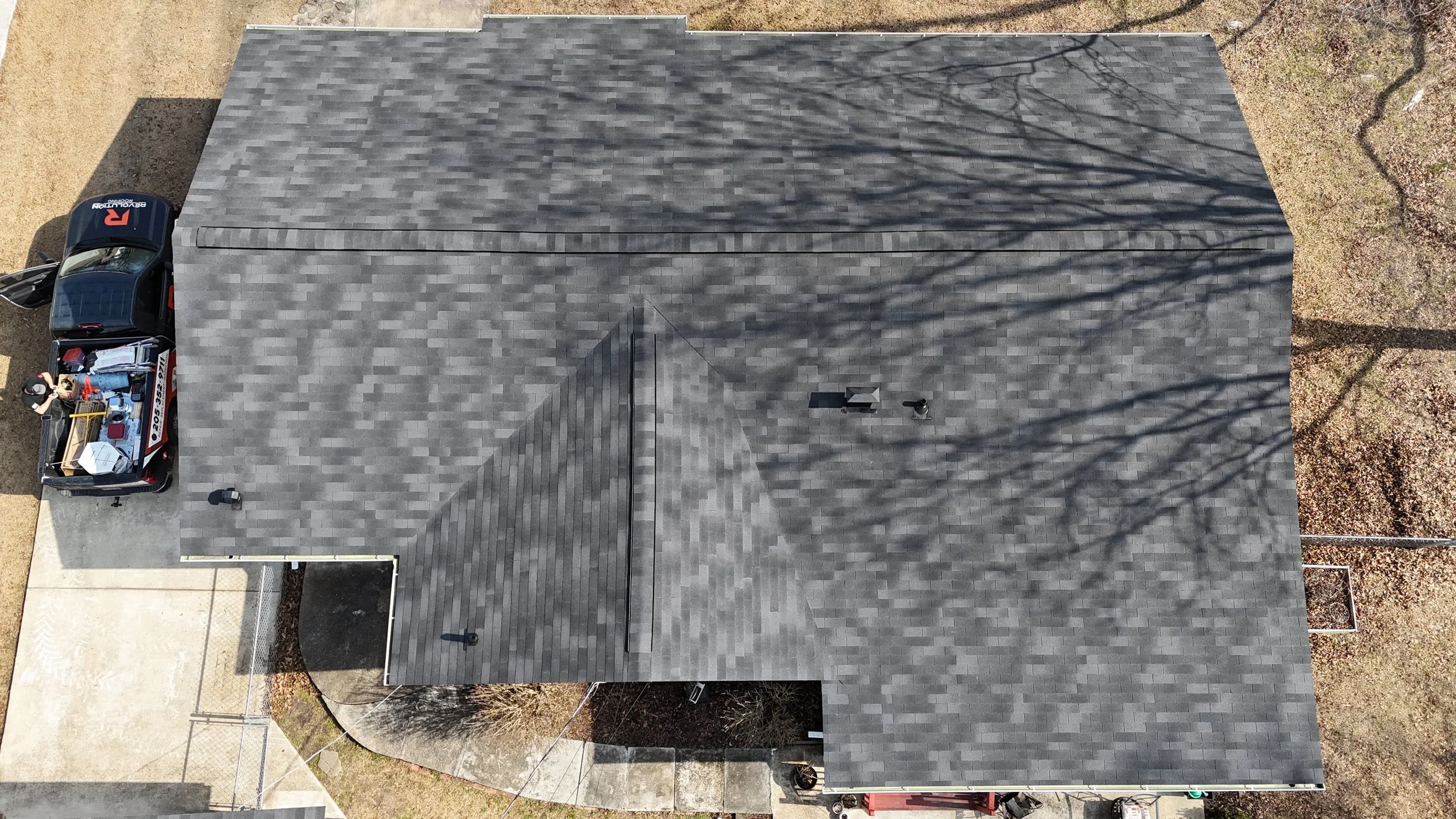 Aerial view of a house with grey shingle roof, a black pickup truck with tools and equipment in the back, and a concrete driveway surrounded by grass and bare trees.