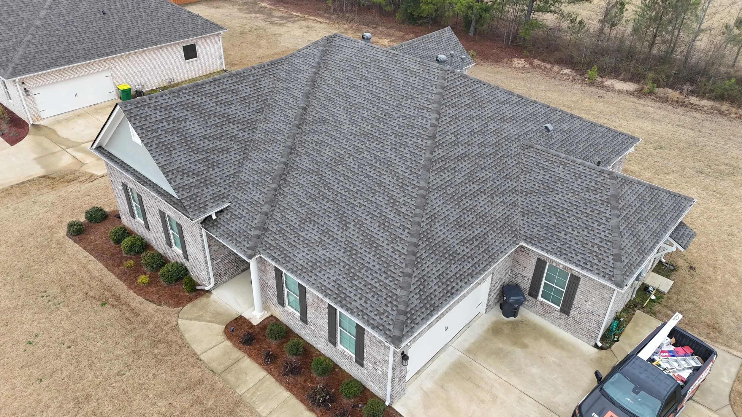 An aerial view of a house with a gray shingled roof, brick exterior, and manicured bushes. Adjacent to the house is a driveway with a vehicle and a neighborhood with other houses and trees.