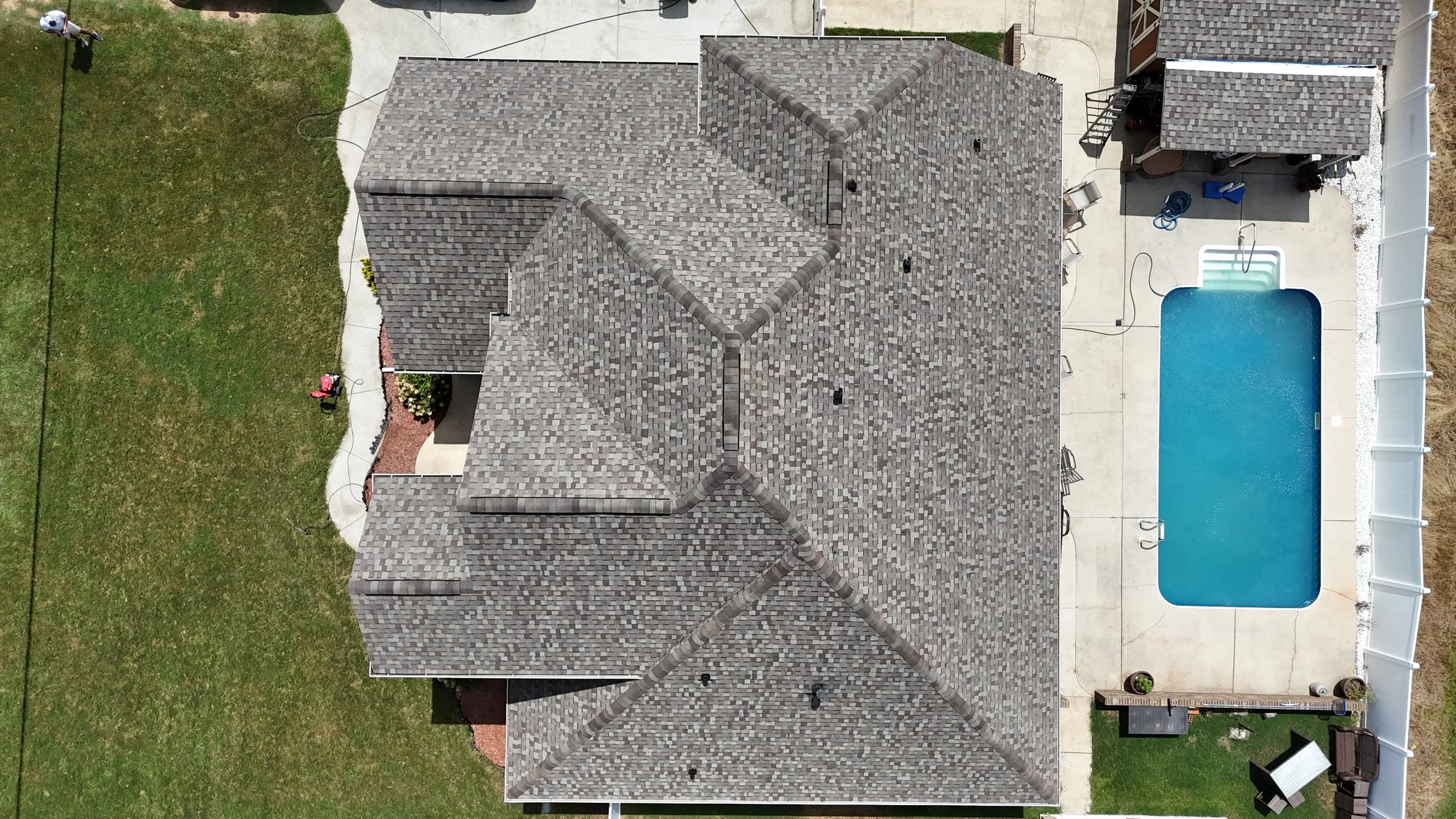 Aerial view of a house with a gray shingled roof, backyard with a pool, patio furniture, and a grassy area with a person watering plants.