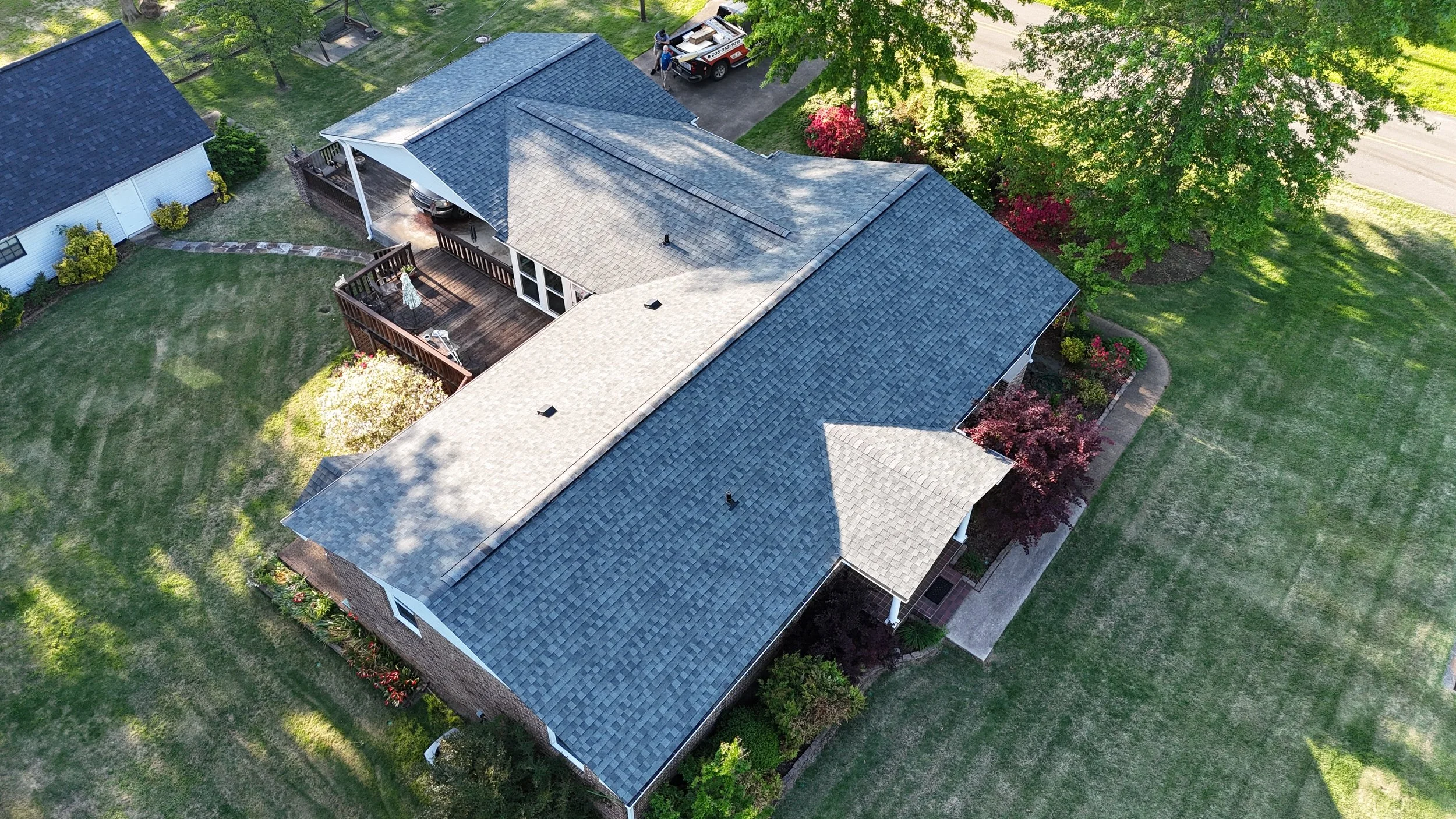 Aerial view of a house with a large yard, trees, a deck, and a trailer parked nearby.