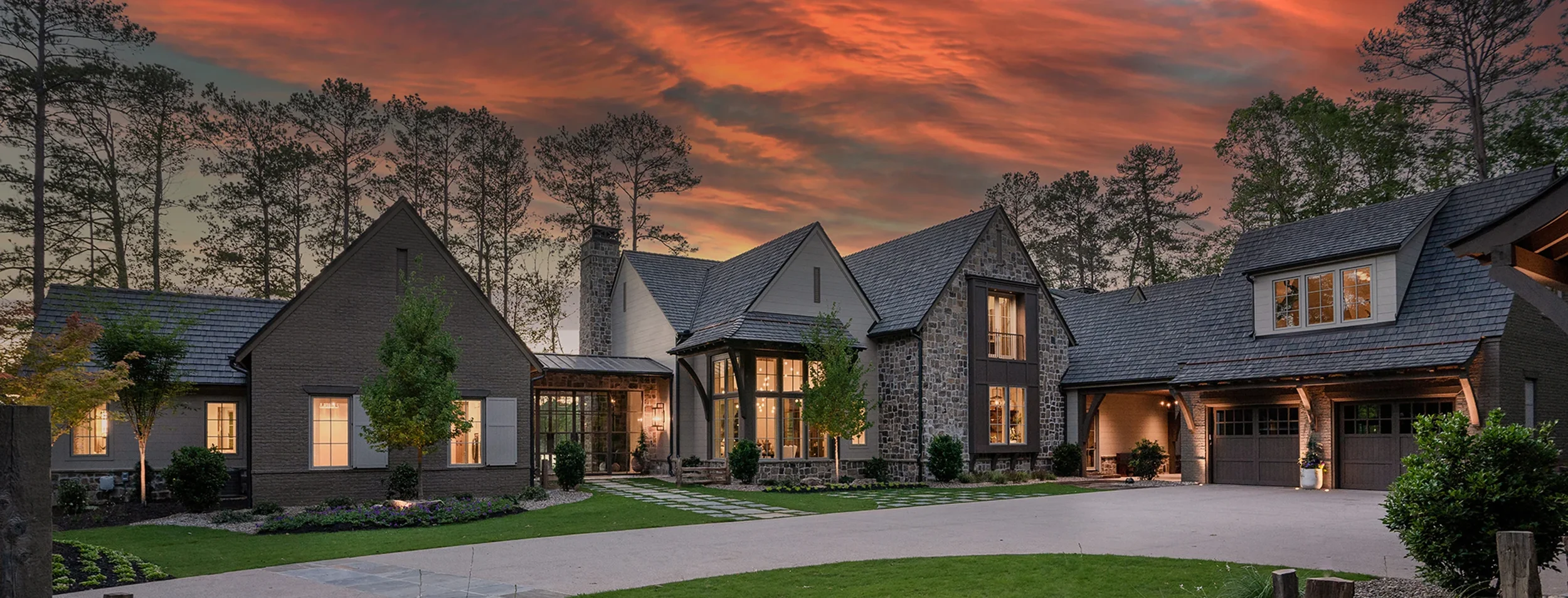 A large, modern house exterior at sunset with a stone and brick facade, large windows, and an attached garage. Surrounding the house are trees, well-maintained landscaping, and a driveway.