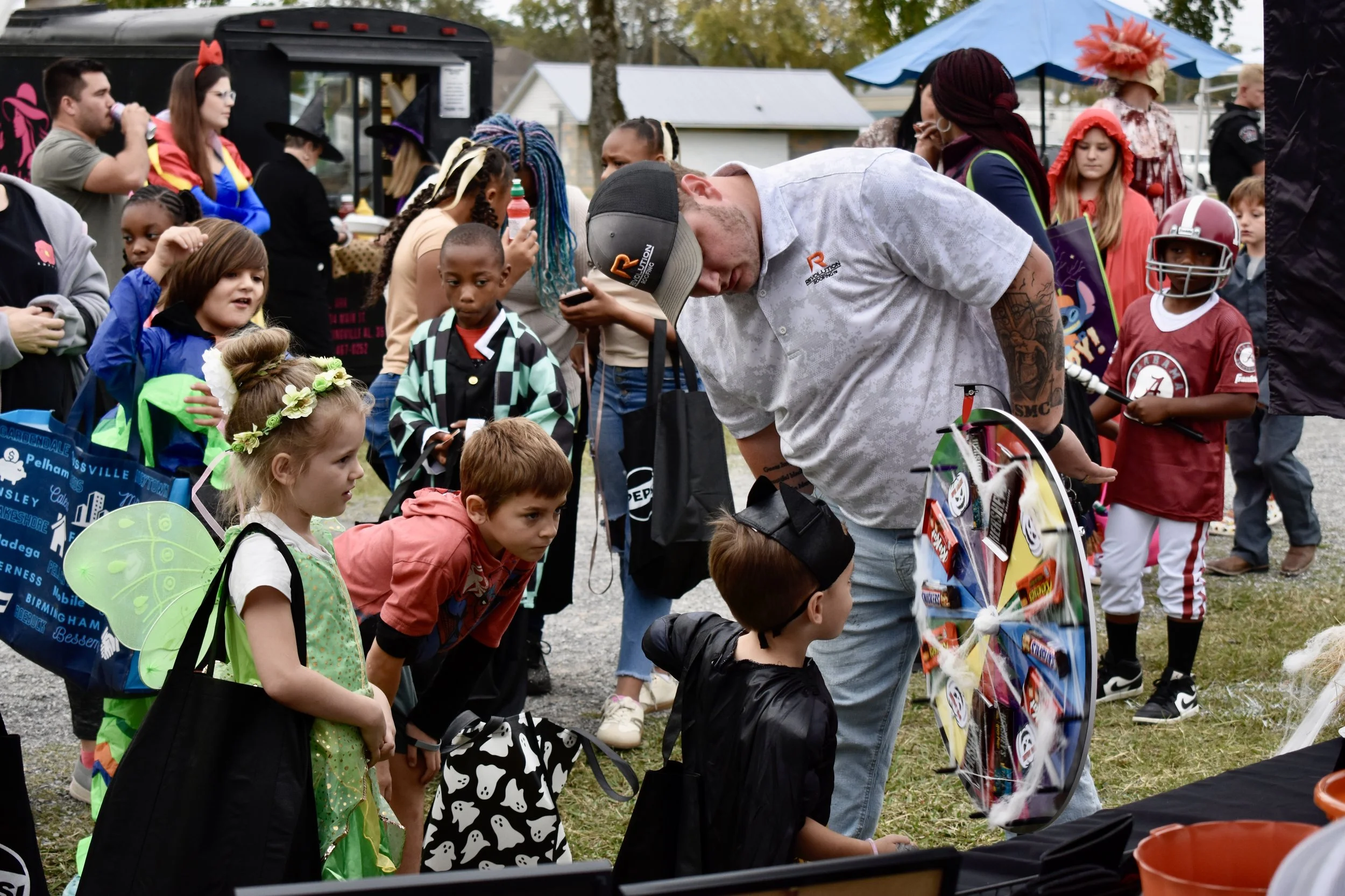 Children and adults at an outdoor event, some dressed in costumes like witches and fairies, gathered around a prize wheel, with booths and trees in the background.