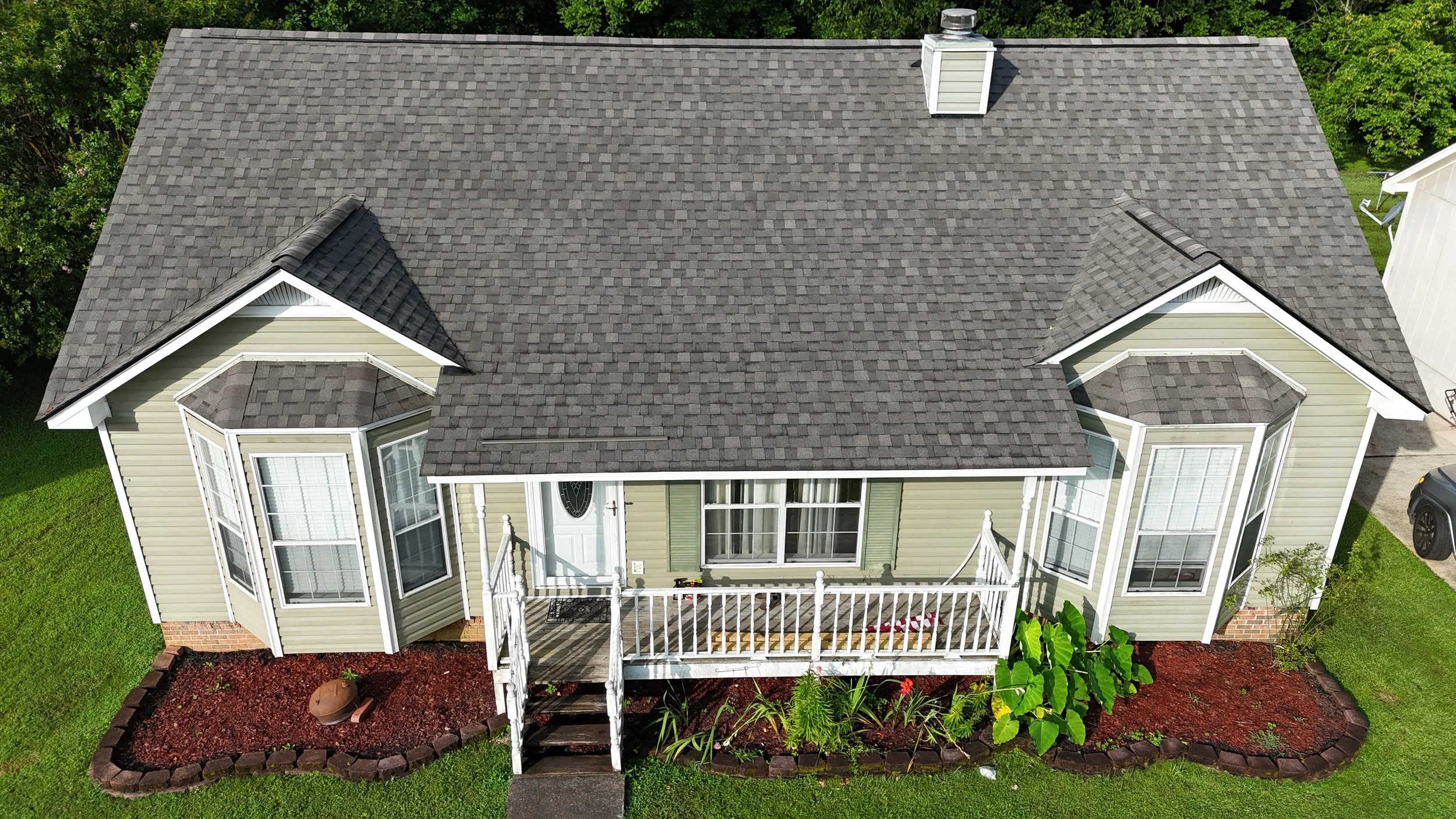 Aerial view of a green and beige house with a gray shingled roof, a front porch with white railings, and a landscaped garden with red mulch and green plants.