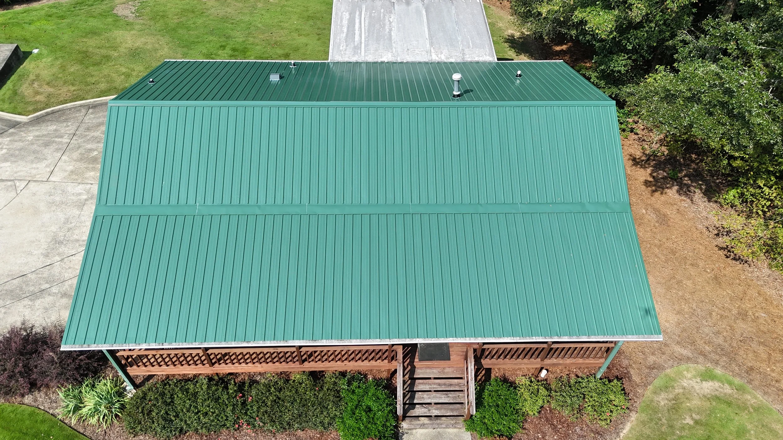 Aerial view of a building with a green metal roof, surrounded by grass, trees, and concrete pavement.