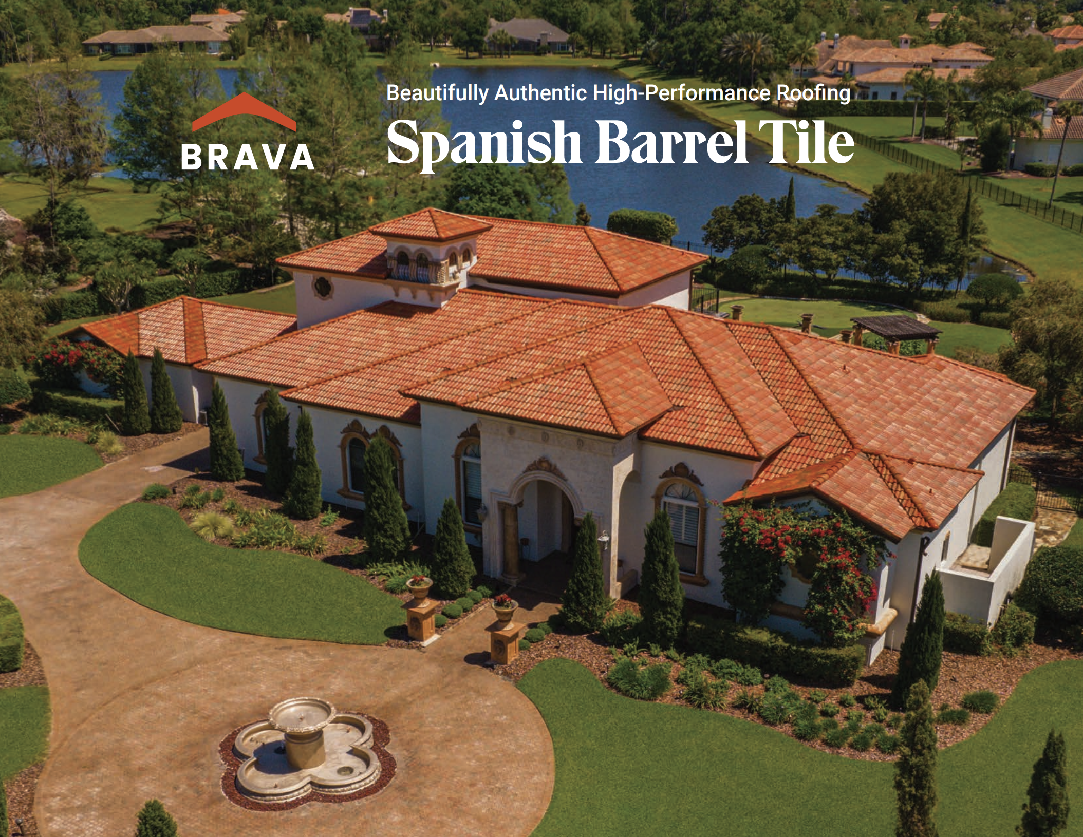 Aerial view of a large house with red tile roof, surrounded by green lawn, trees, and shrubs, with a pond or lake nearby and a fountain in the front yard.