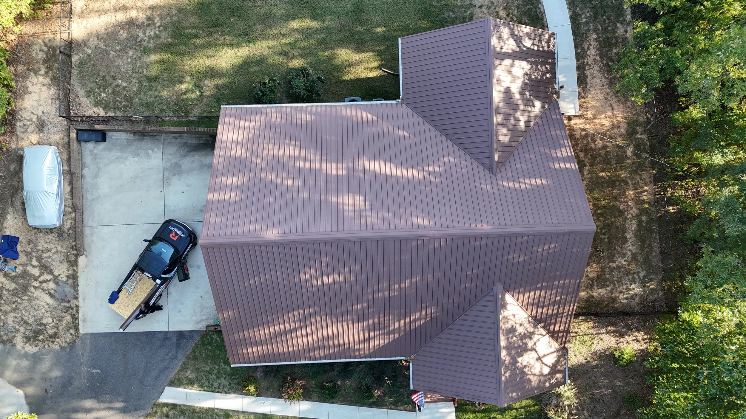 Aerial view of a house with a rusty red metal roof, surrounded by green trees and a yard. There is a driveway with a covered car, a trailer, and a few items near the house.