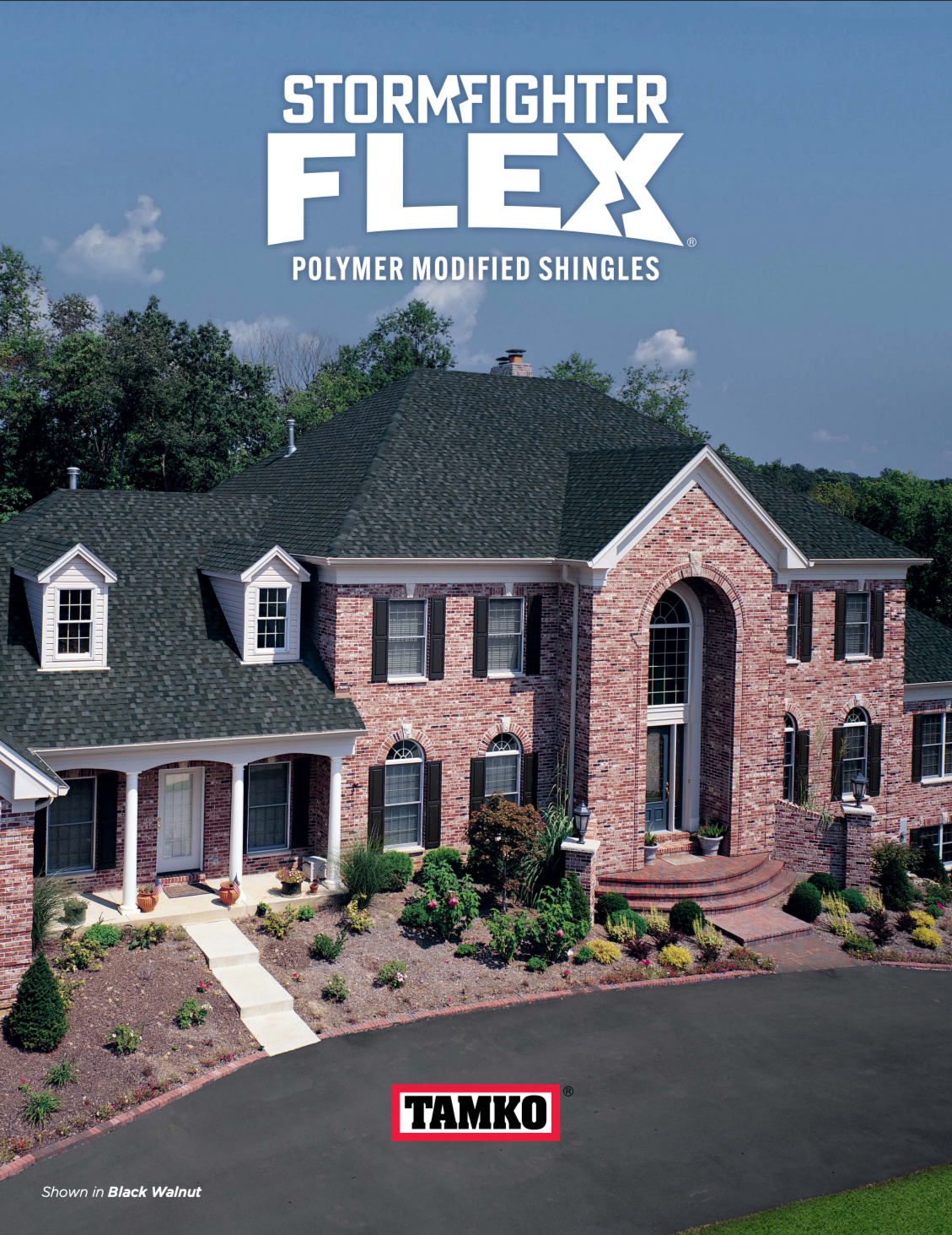 A large brick house with a dark shingle roof, multiple windows with black shutters, and a front porch with white columns. The house has a landscaped yard with bushes and flowers, set against a backdrop of trees and a blue sky with some clouds.