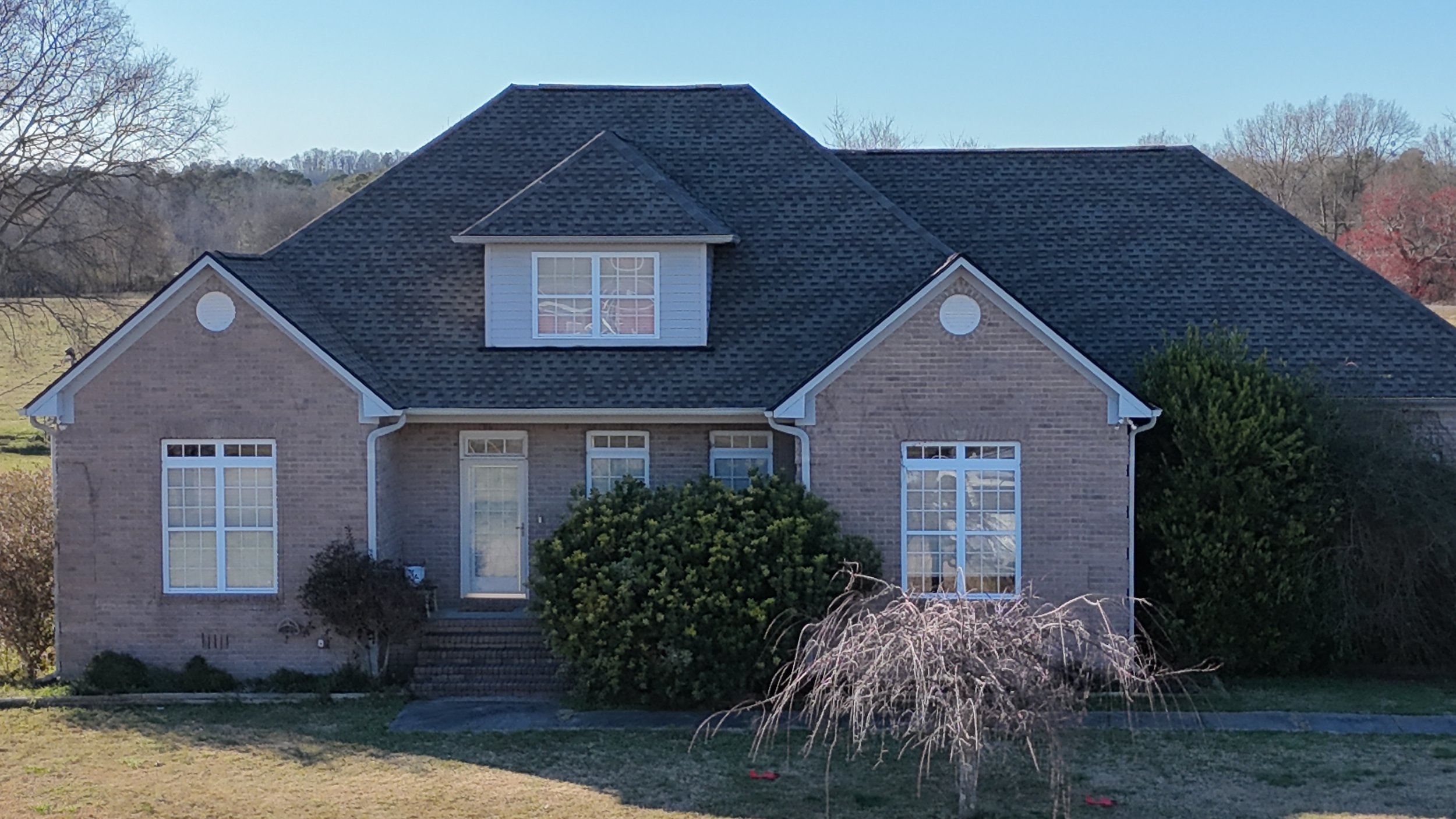 Two-story brick house with a dark shingled roof, large windows, and a front porch. There are shrubs and bushes in front of the house, with a grassy yard and leafless trees in the background during daylight.