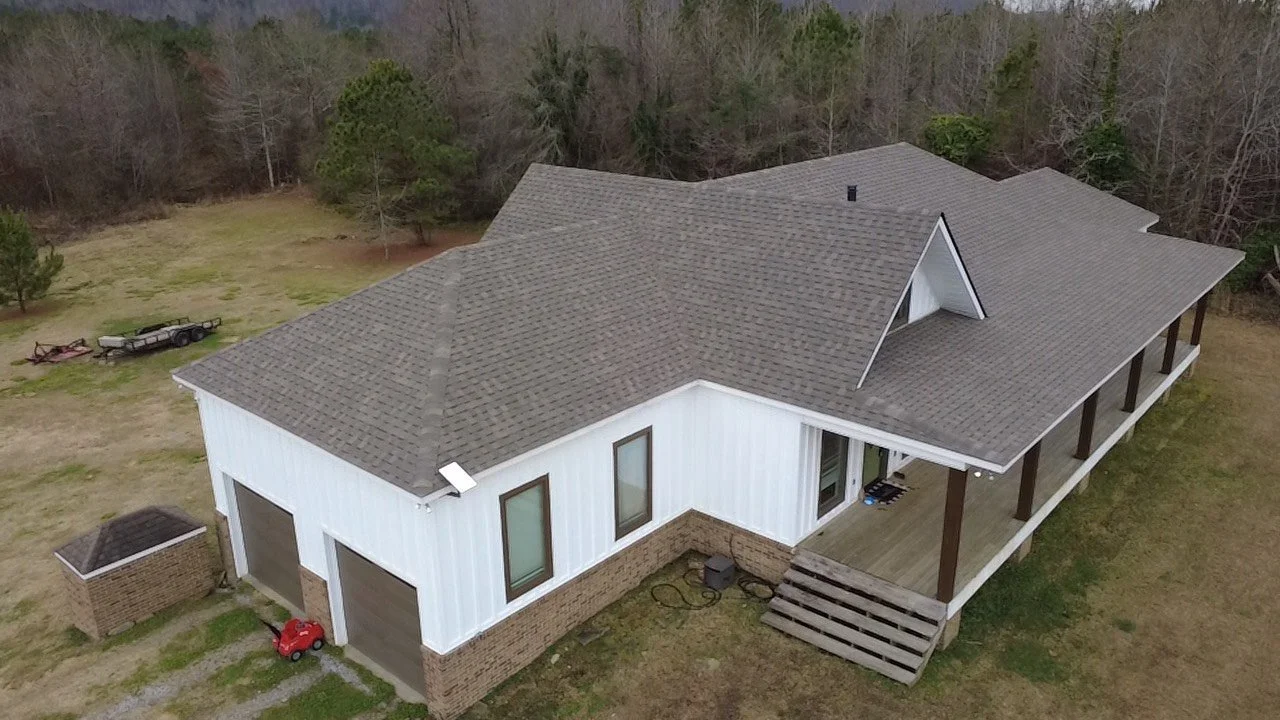 An aerial view of a two-story house with a gray asphalt shingle roof, white siding, and a brick foundation. There are two garage doors on the side, and a small red toy car is parked outside. The house features a porch with wooden support beams and steps leading up to it. The surrounding area has some trees and an open yard.