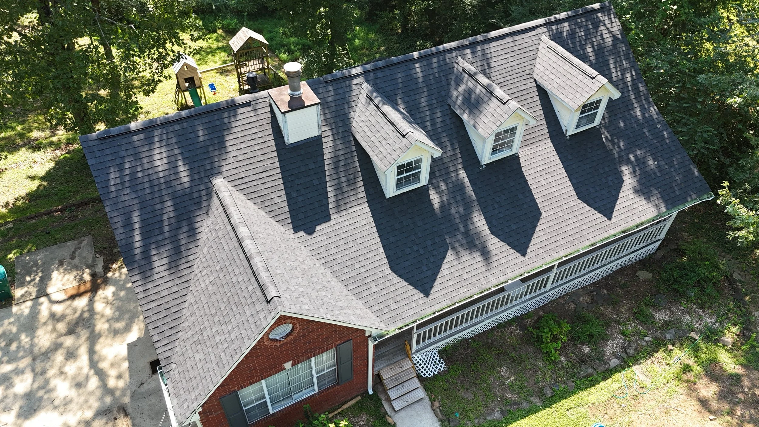An aerial view of a house with a gray shingled roof and three dormer windows. The house has a brick chimney, a white railing porch, and a backyard with a small playground surrounded by trees.
