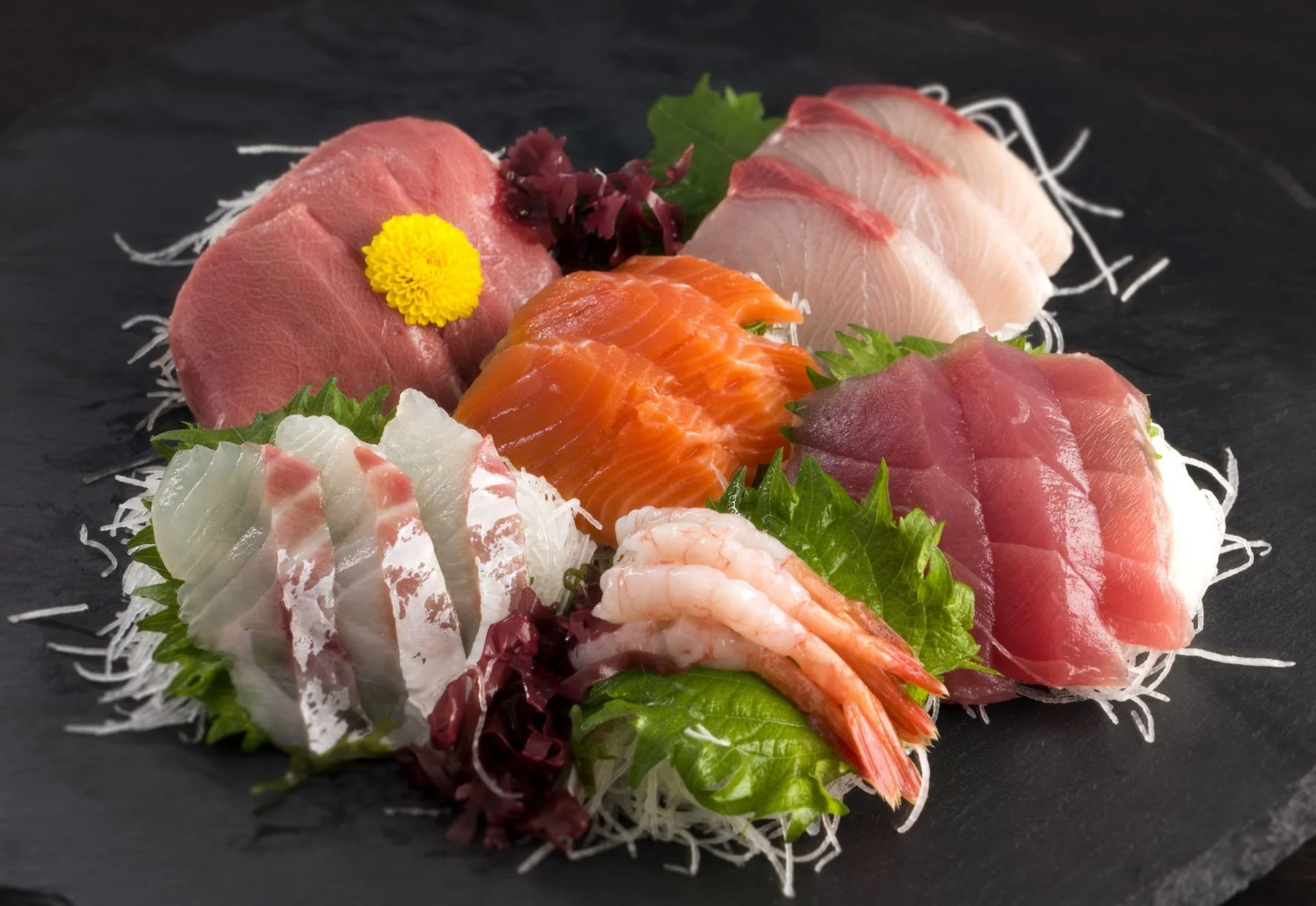 Assorted slices of raw sashimi, including tuna, salmon, yellowtail, shrimp, and white fish, garnished with green leaves, yellow flower, and purple seaweed on a black slate plate with shredded daikon radish.