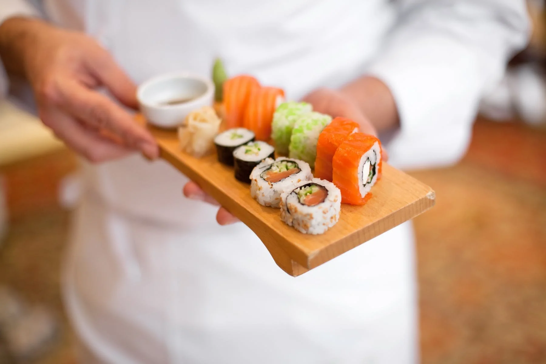 Person holding a wooden platter with assorted sushi rolls, including salmon, cucumber, and avocado, accompanied by a small dish of soy sauce.