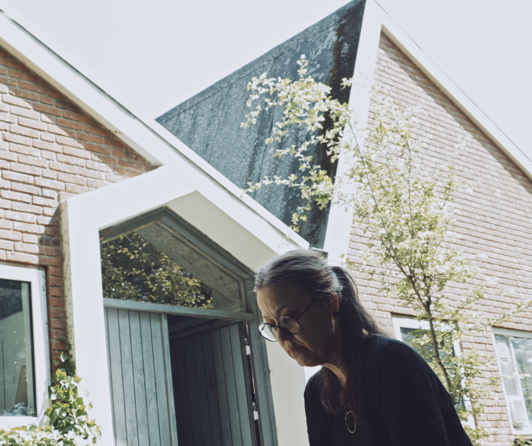 Trine Boesen foran sit atelier i dagslys, omgivet af arkitektur, træer og grønne omgivelser.