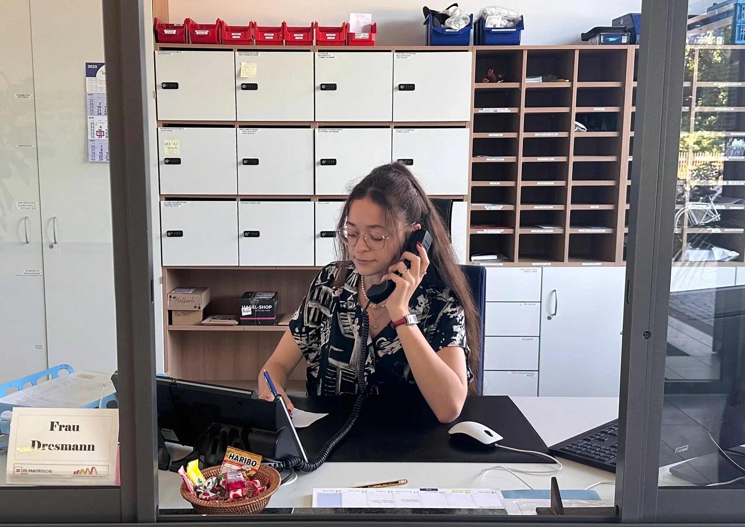 A woman with dark hair, glasses, and a patterned blouse is sitting at a desk in an office, talking on the phone, with a computer monitor, a keyboard, and a mouse in front of her. Behind her are white lockers, open shelves with various items, and some window showing trees outside.