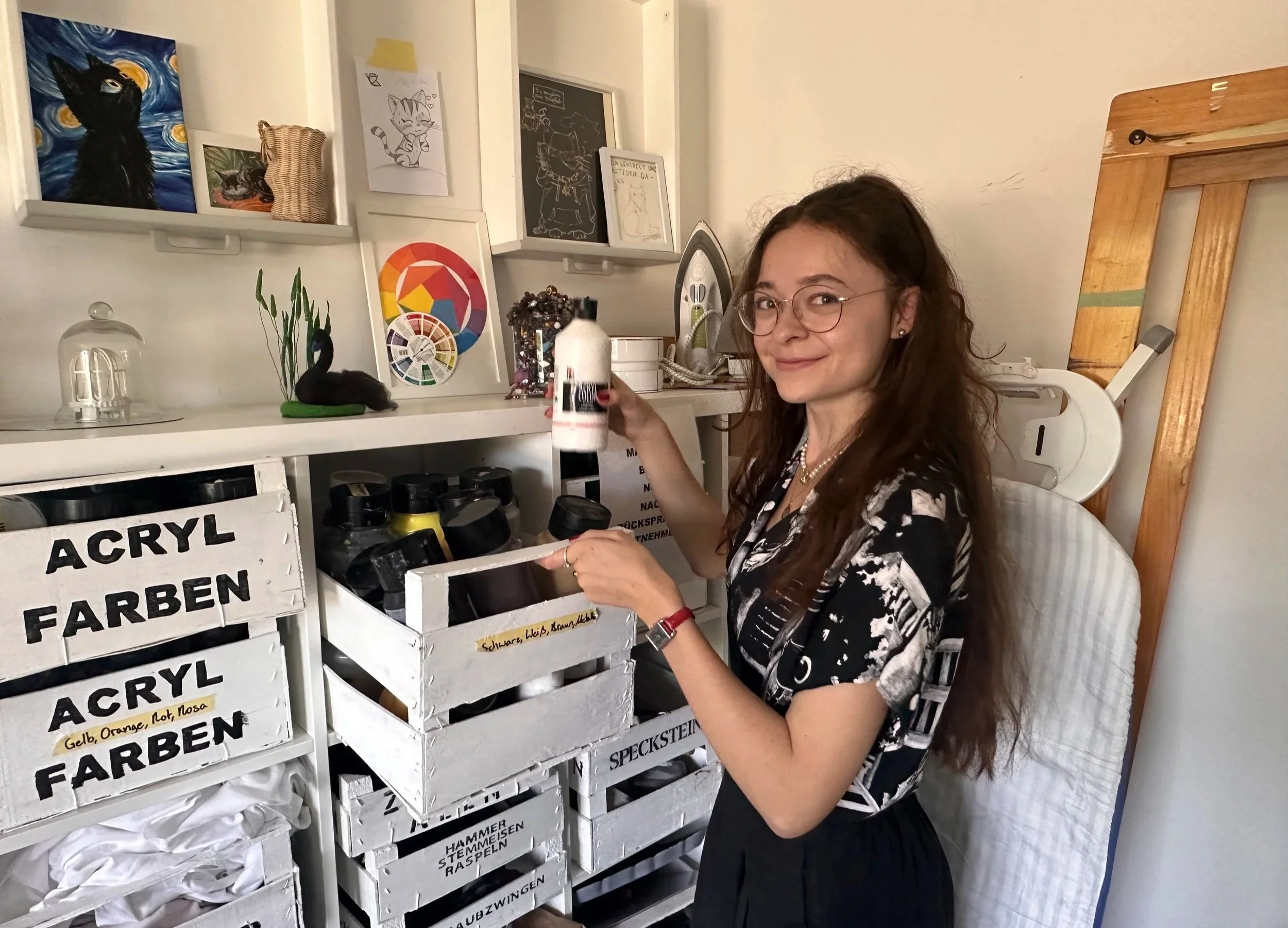 A woman with glasses and long hair smiling at the camera, standing next to a white shelf with labeled bins filled with colored pencils. The room has artwork on the wall, including paintings and drawings of cats, an iron, a plant, and a wooden mirror leaning against the wall.