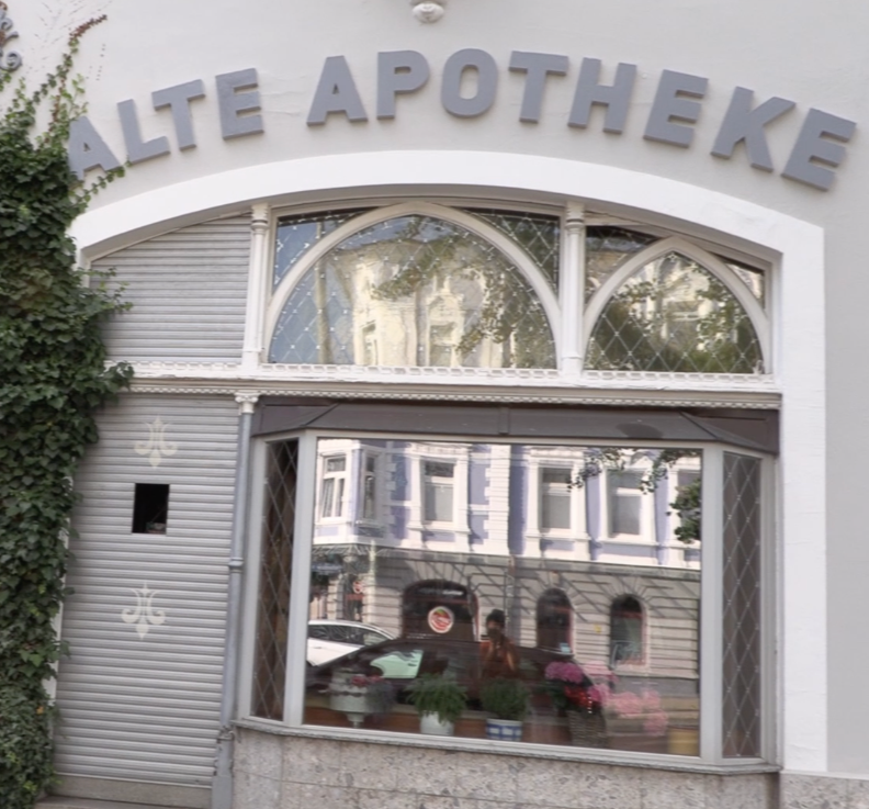 The storefront of a pharmacy with a sign in German reading 'Alte Apotheken', featuring a large arched window, surrounded by white walls and partially covered with green ivy.